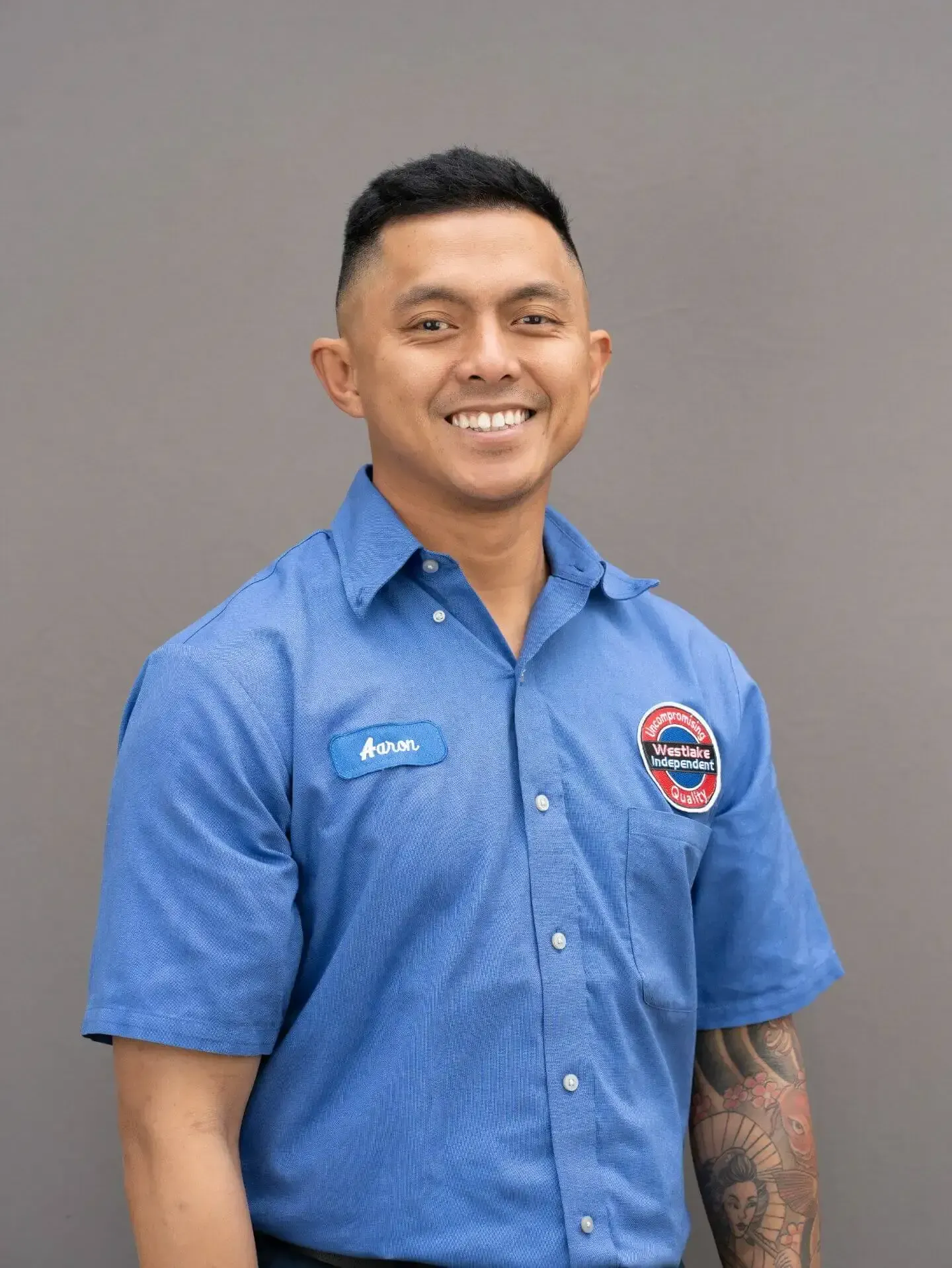 Smiling person in a blue button-up shirt with a logo, standing against a plain gray background | Westlake Independent Automotive