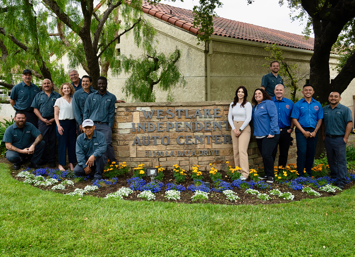 Group of people posing by a stone sign in a landscaped lawn with purple flowers and trees. | Westlake Independent Automotive