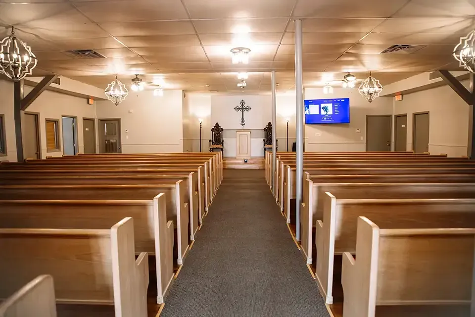 Chapel with wooden pews, central aisle, podium, cross, and a blue display screen inside