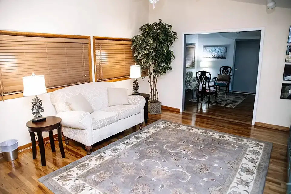 Bright sitting room with white sofa, lamps, large rug, and additional seating beyond open doorway