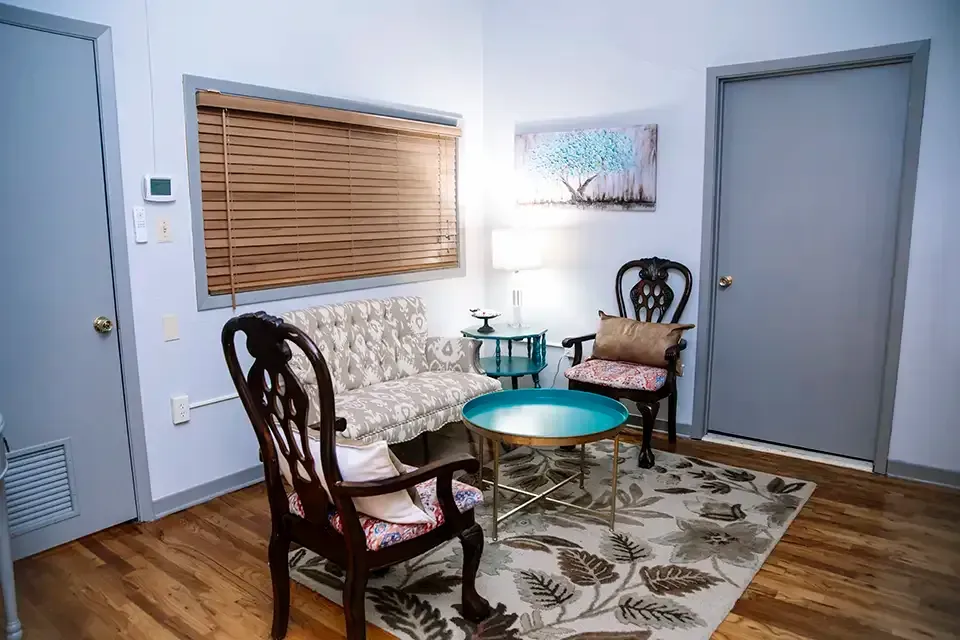 Small sitting room with floral sofa, two chairs, round blue table, rug, lamp, and closed blinds and doors
