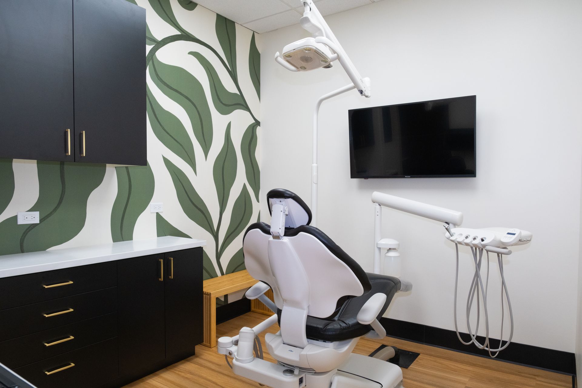 Dental exam room with chair, tools, mounted TV, cabinets, and leaf-patterned wall.