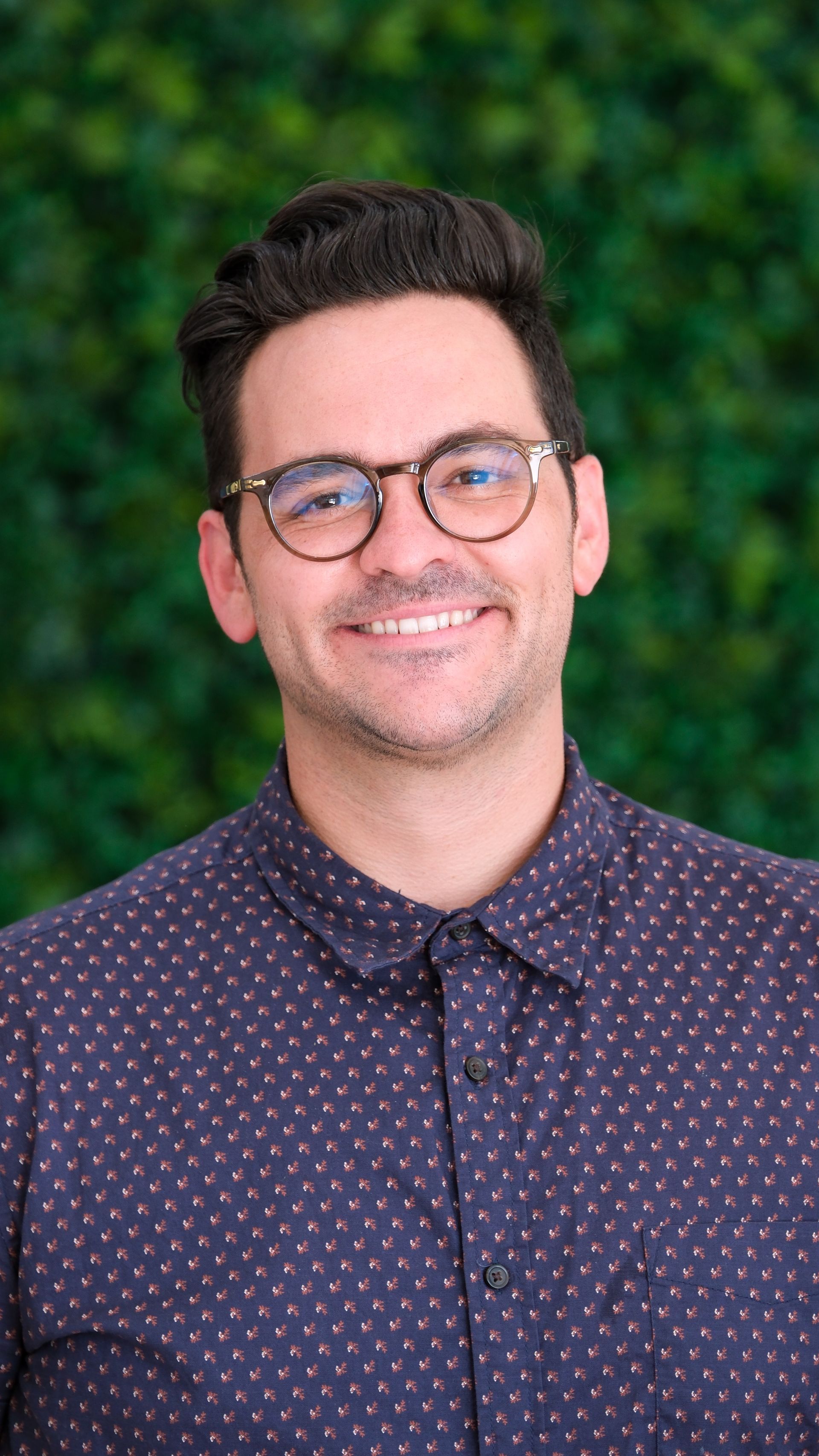 Man wearing glasses smiles, standing in front of a green wall, wearing a navy button-down shirt with white dots.