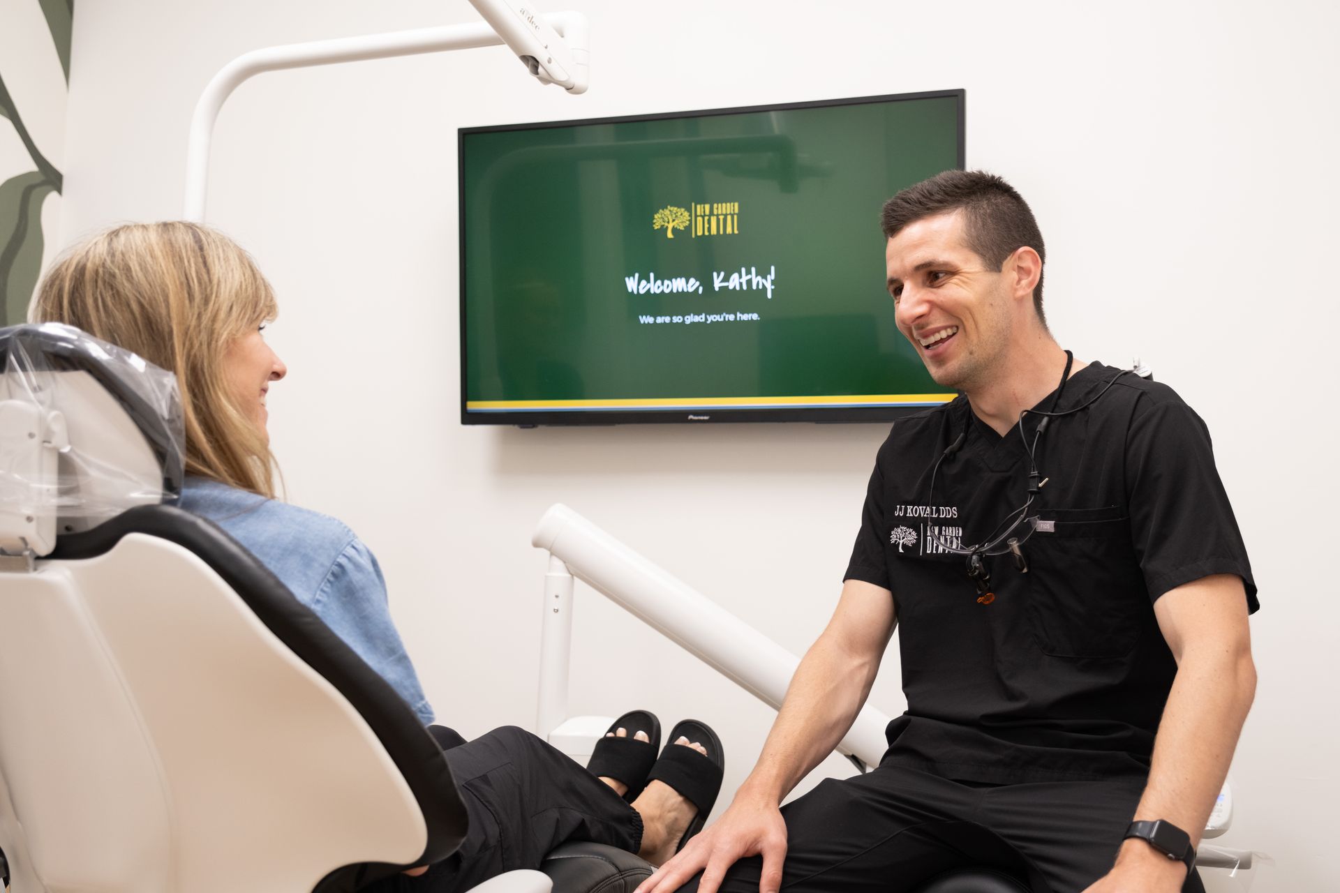 Dentist talking with a patient in a dental chair. Office setting, TV screen on the wall.