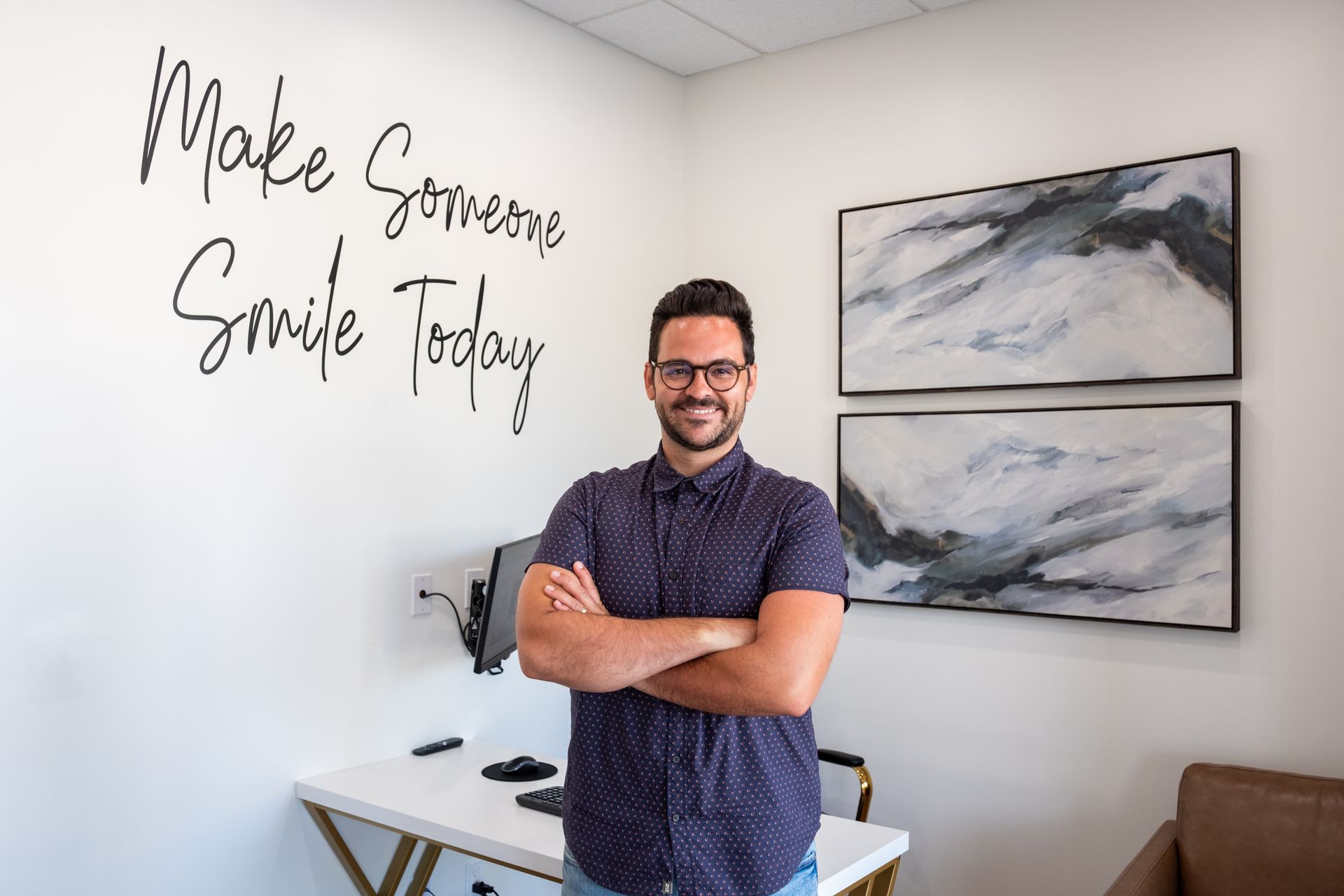 Man with arms crossed, smiling, standing in a brightly lit office. White walls, art, desk, text