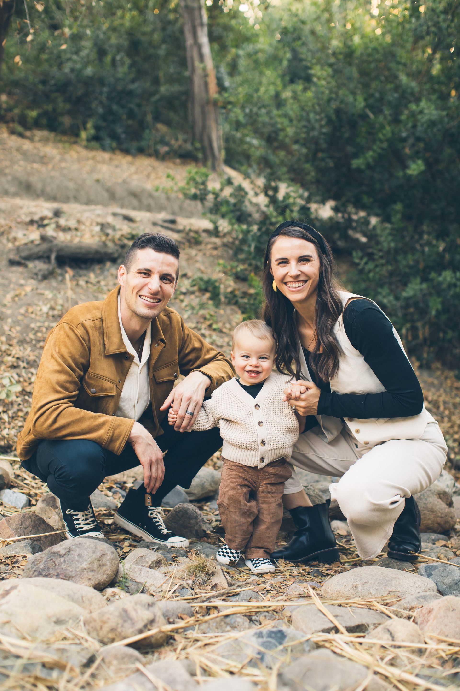 Family poses outdoors, smiling. Man in brown jacket, woman in vest, toddler in front, standing on rocks.