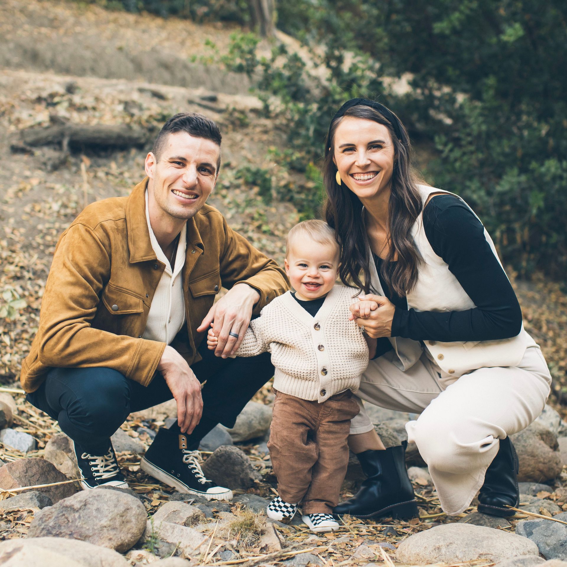 Family poses outdoors, smiling. Man in brown jacket, woman in vest, toddler in front, standing on rocks.