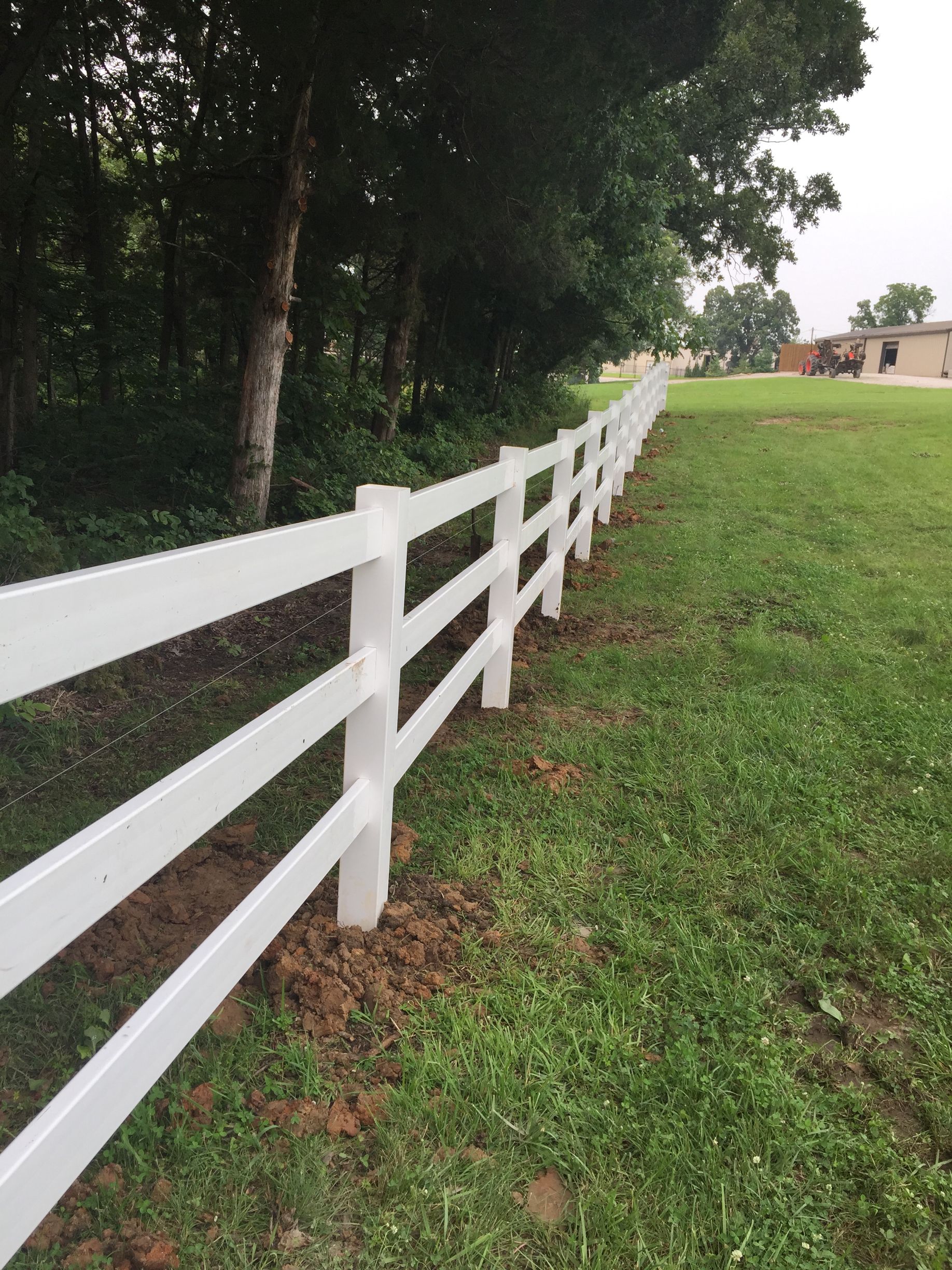 White three-rail fence alongside a grassy field, with trees in the background.