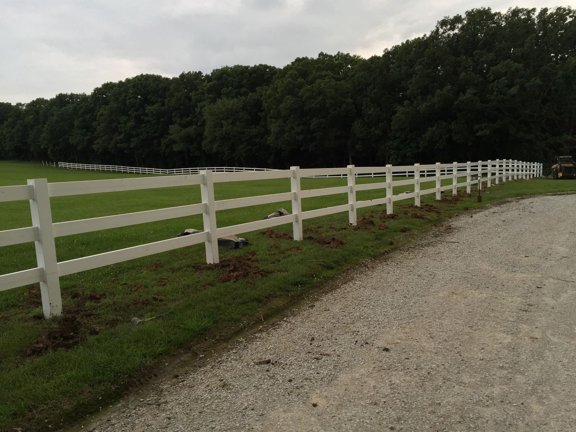 White three-rail fence alongside a gravel driveway and green lawn, with trees in the background under an overcast sky.
