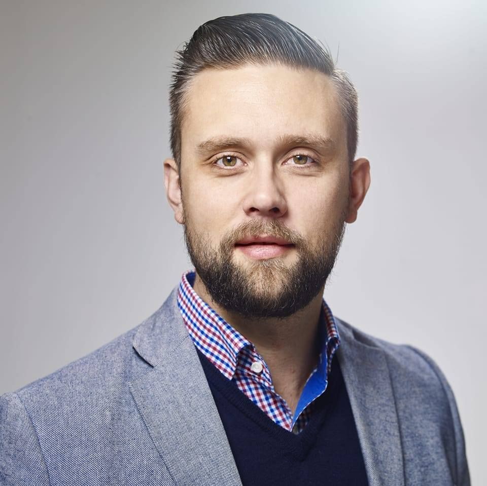Man with a beard, wearing a blazer over a collared shirt and sweater, looking at the camera.
