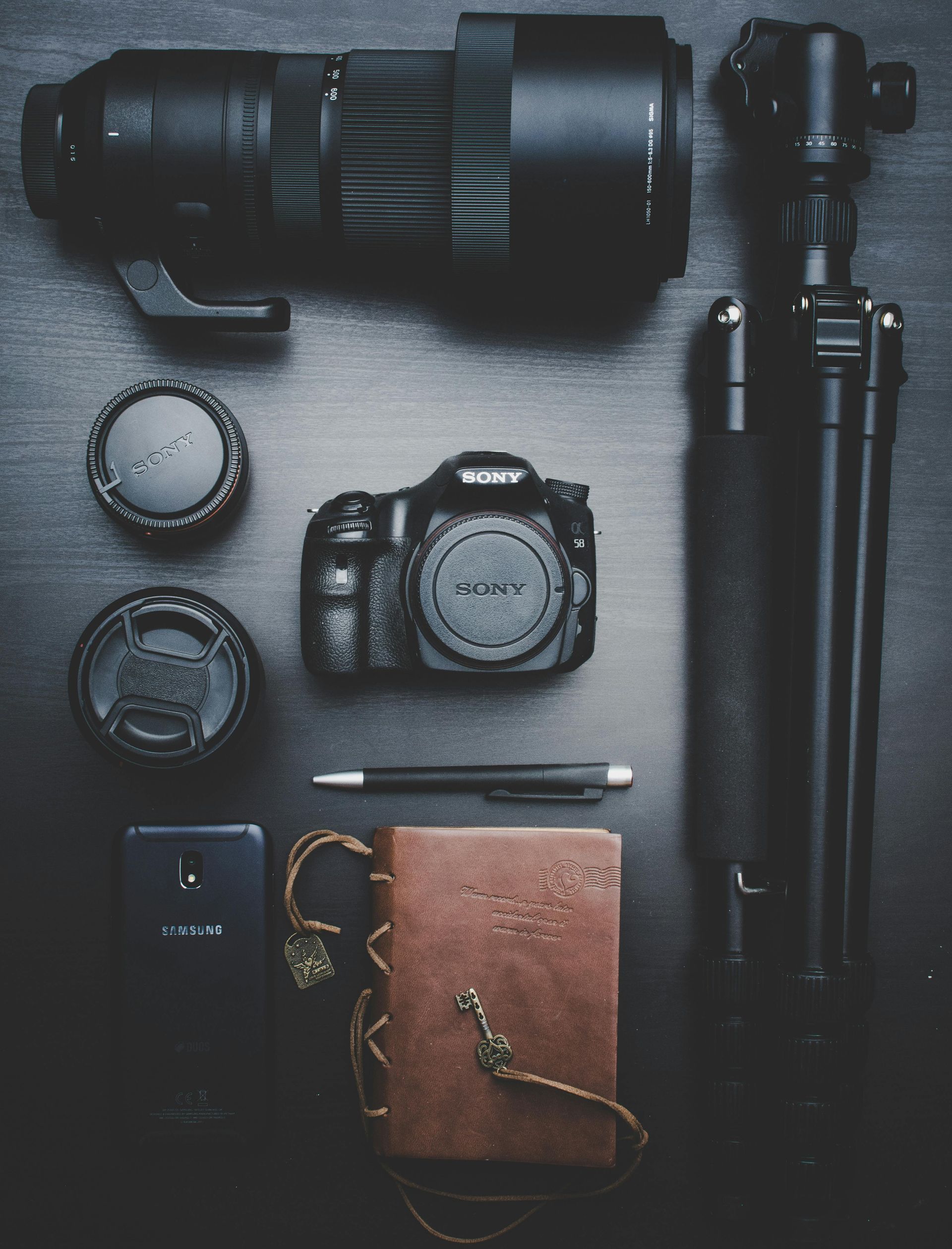 Camera equipment laid out on a dark surface: camera, lens, tripod, lens caps, journal, pen, phone.