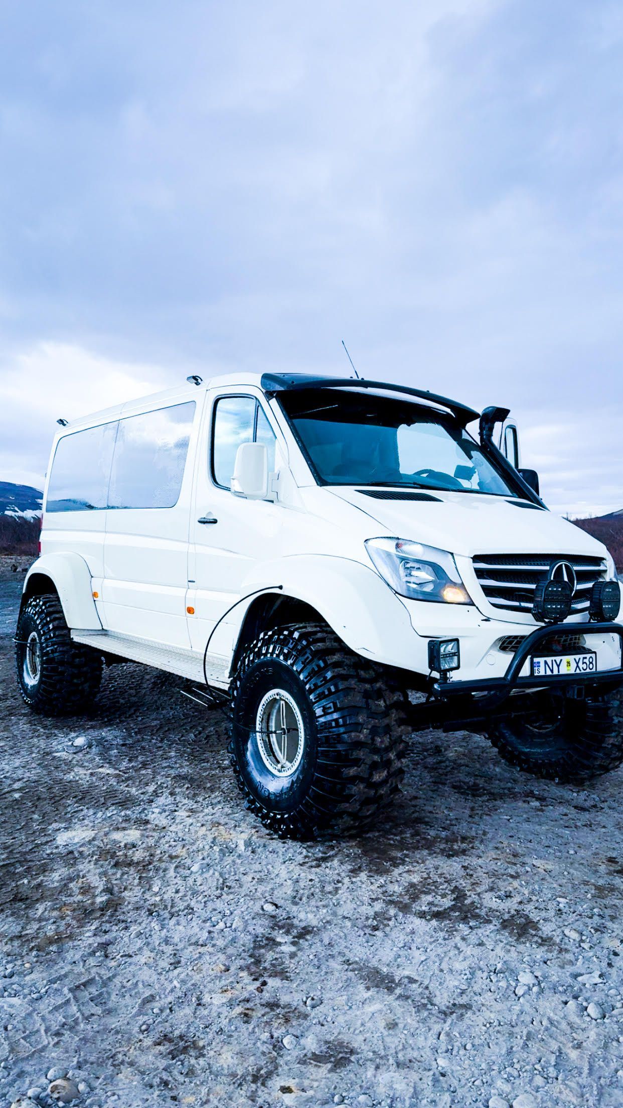 White, off-road van driving in snow. Has large tires and blue logo. Winter setting.