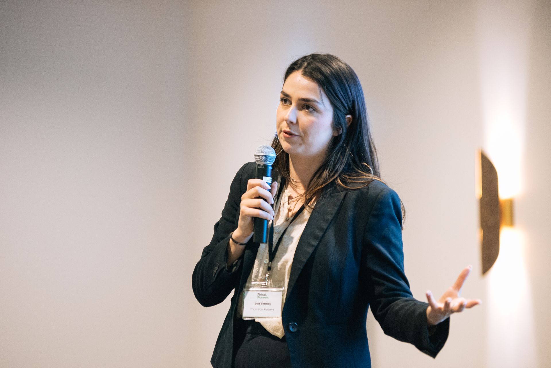 Woman speaking into a microphone at a conference, wearing a dark blazer and gesturing with one hand.