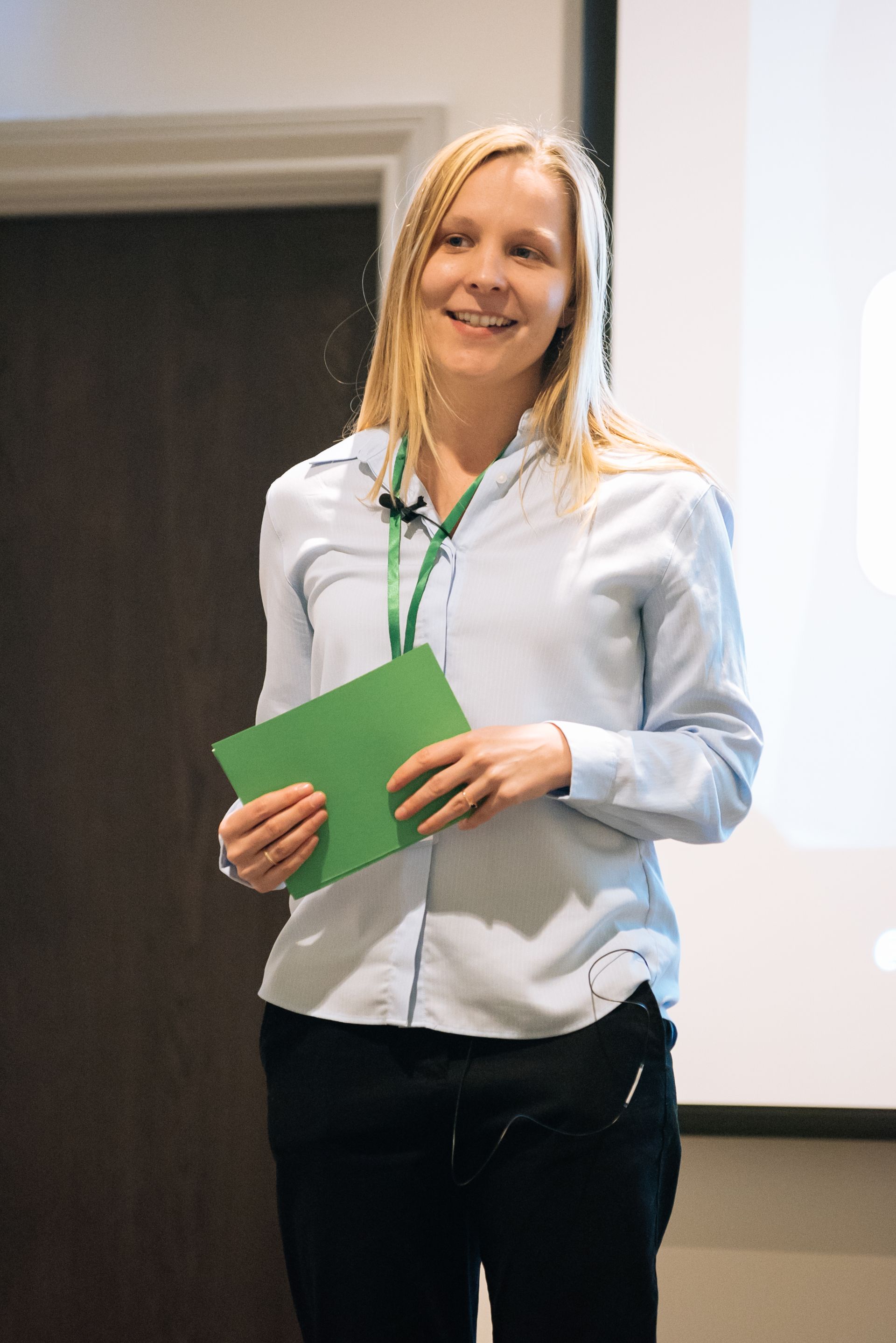 Woman in a white shirt holding a green folder and smiling on a stage or conference room