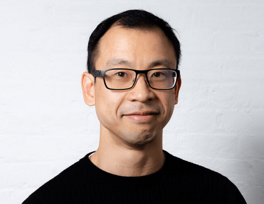 Man with glasses smiling in front of a white brick wall, wearing a black sweater.
