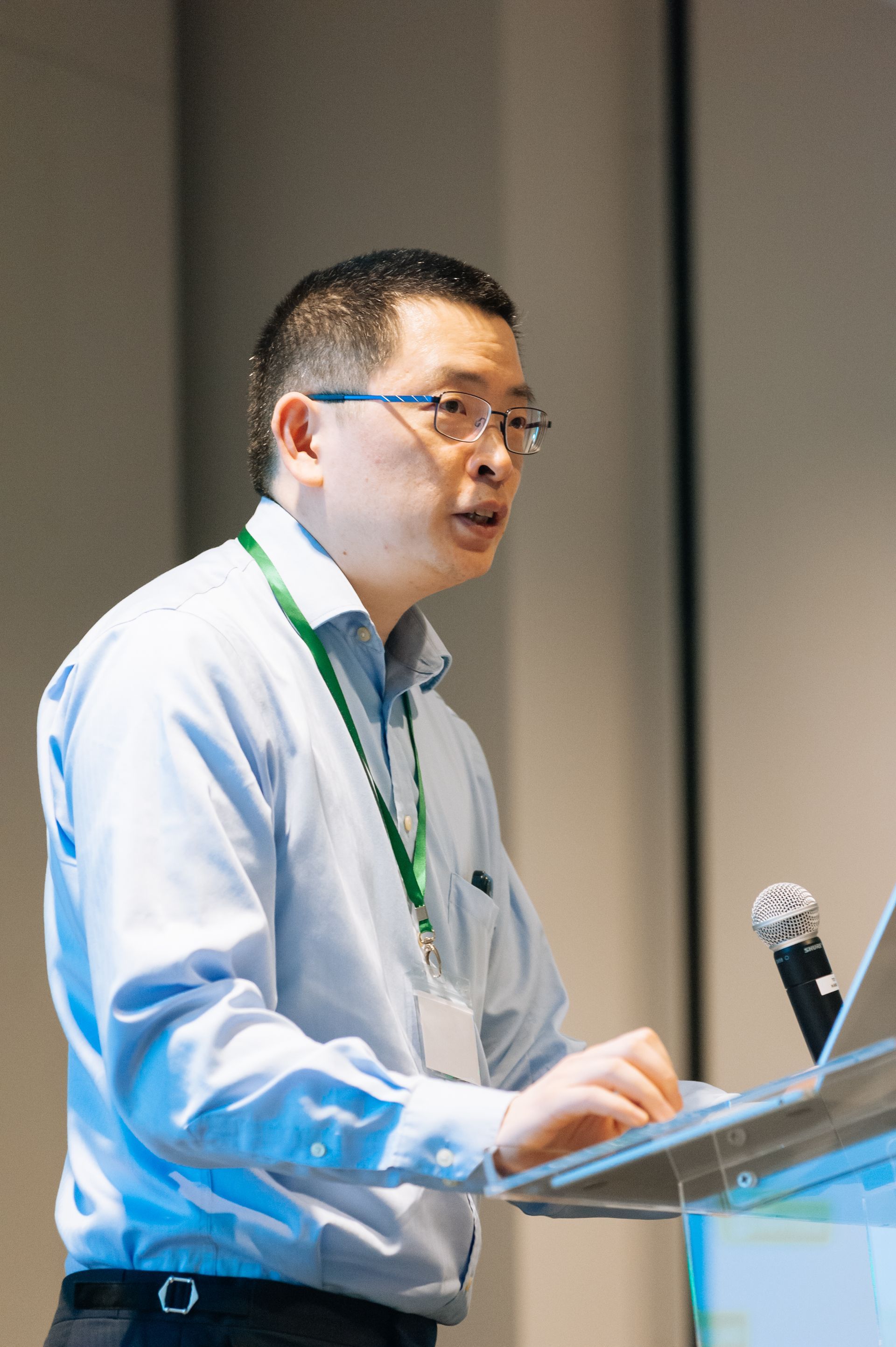 Man speaking at a podium with a microphone, wearing a light blue shirt and green lanyard