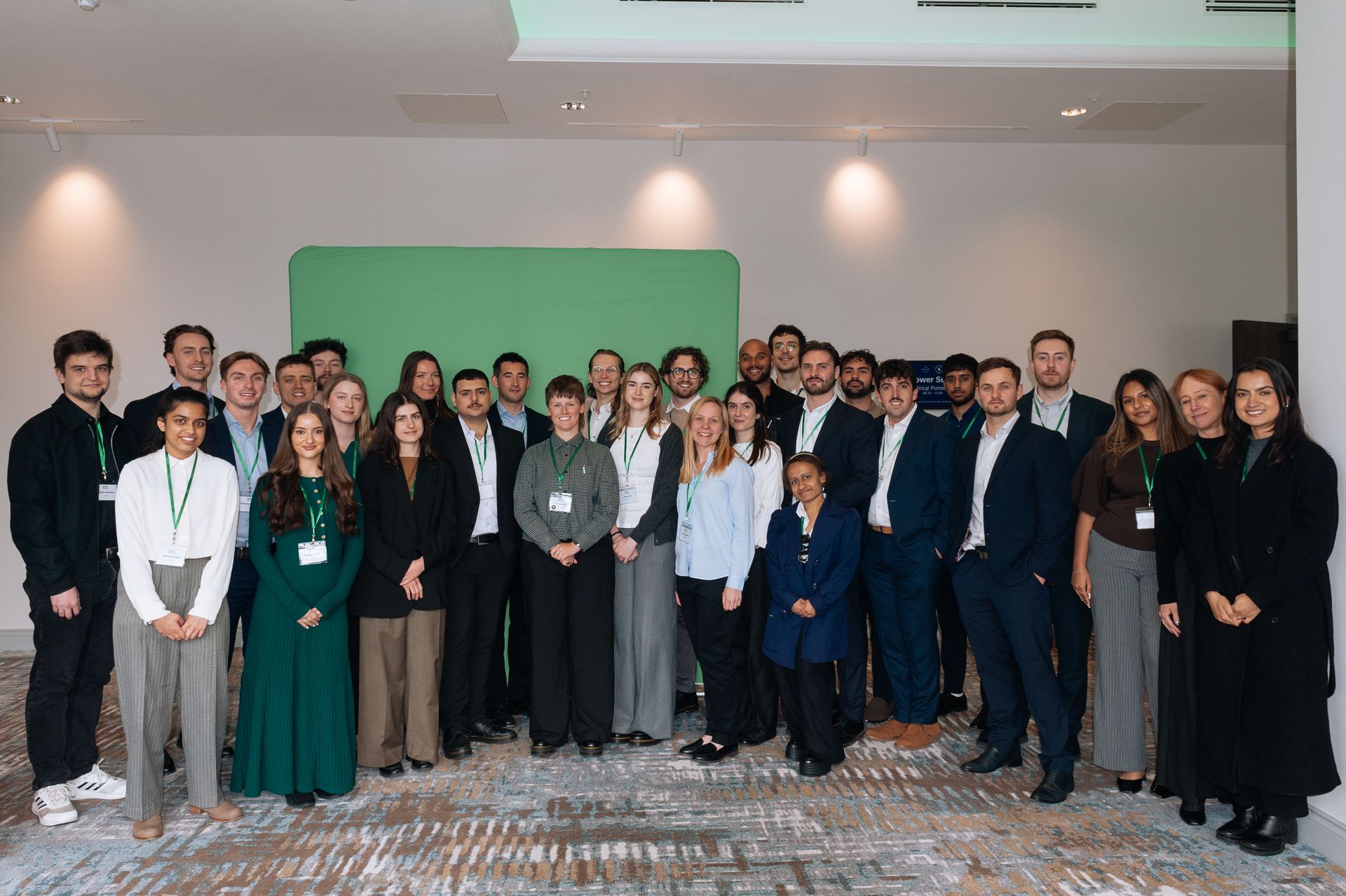Large group posing indoors at a formal event, with a green backdrop and carpeted floor.