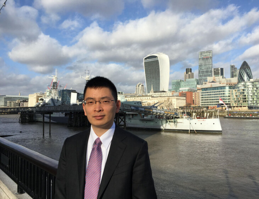 Man in suit poses in front of London skyline with Thames River and HMS Belfast warship on a sunny day.