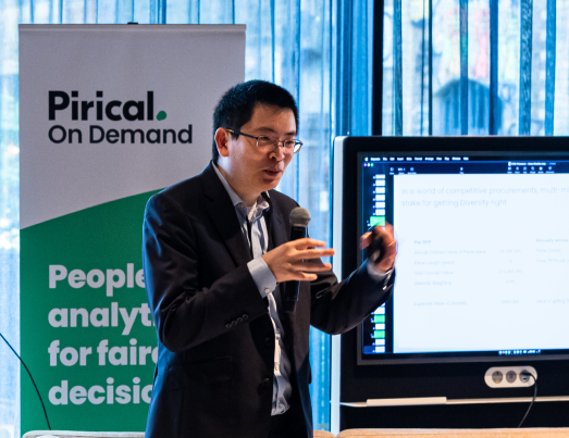 A man in a suit gestures while speaking at a presentation, in front of a monitor and a 