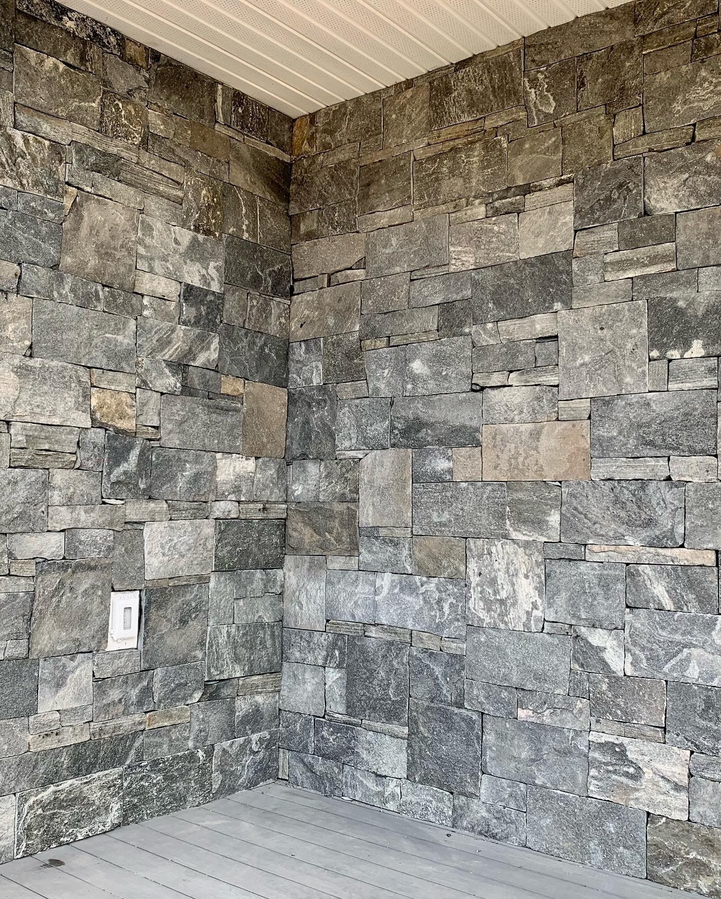 A corner of an outdoor wall made of rectangular, multi-toned gray stone blocks, with a white electrical outlet visible.