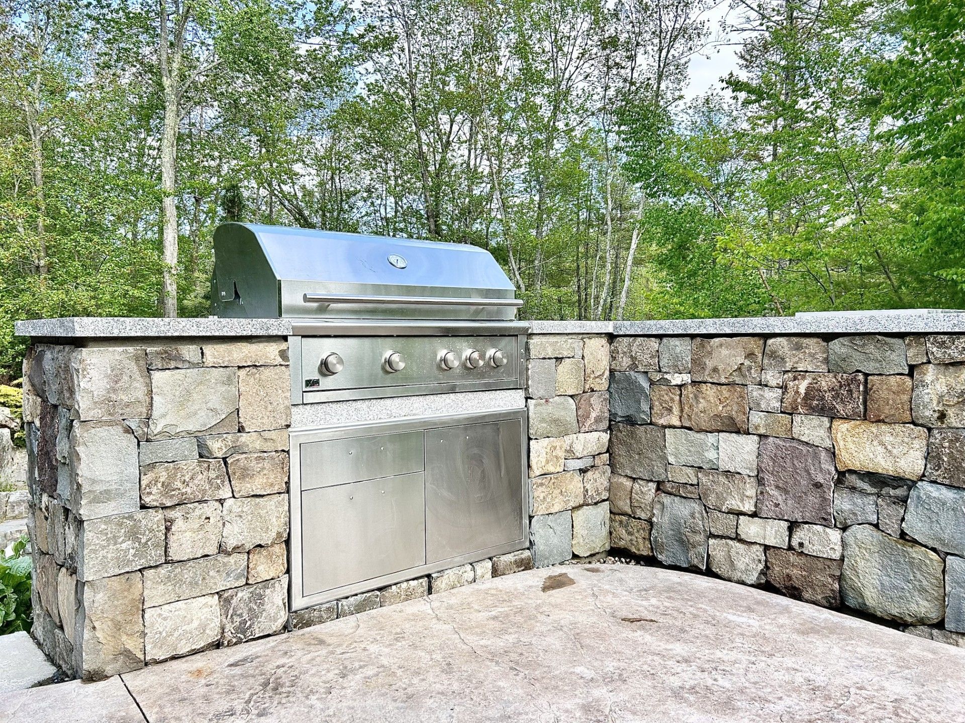 An outdoor stainless steel gas grill built into a rustic stone kitchen island on a patio with a background of green trees.