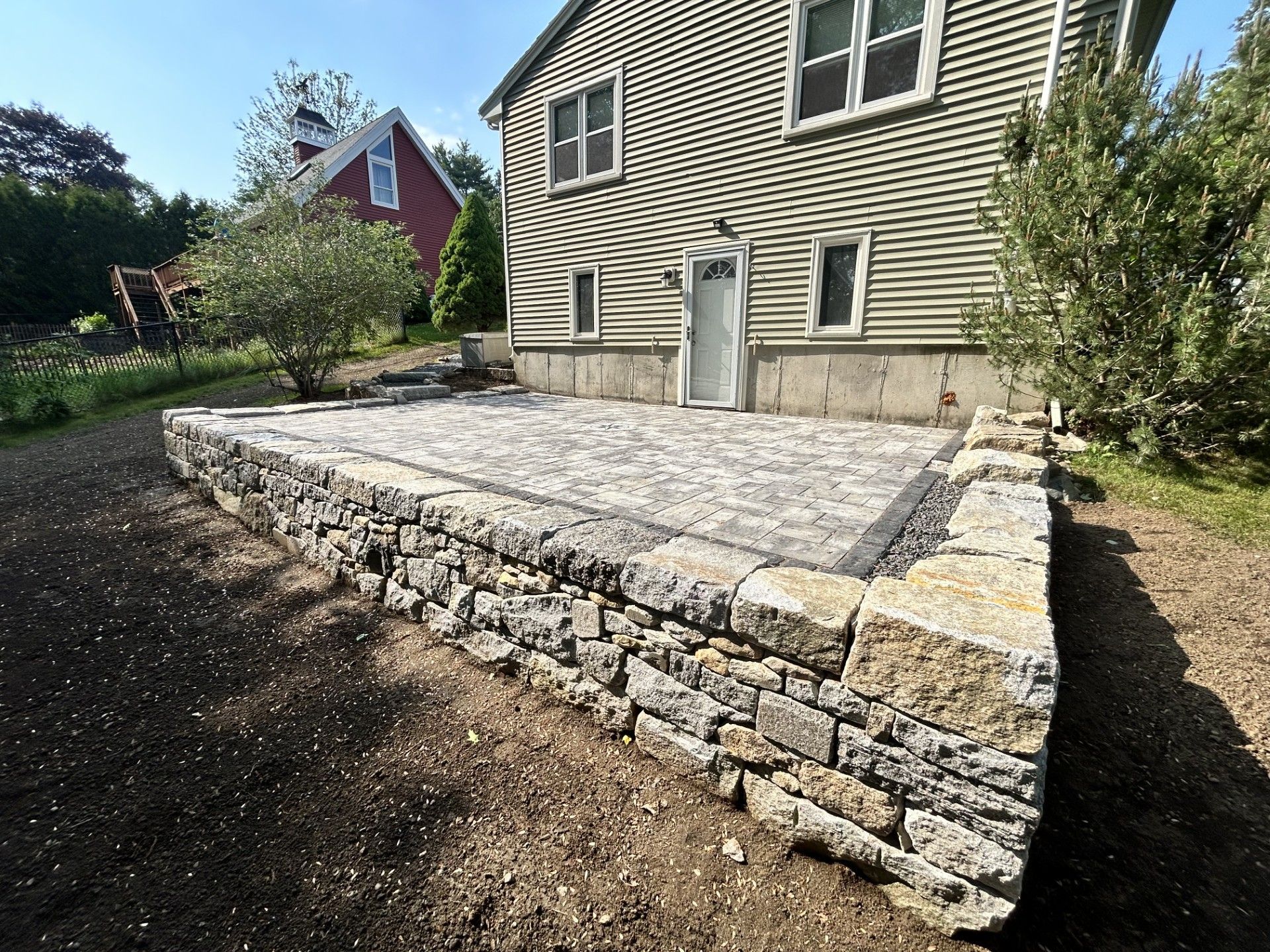 A newly constructed stone retaining wall supporting a paved patio outside a beige house with a red building nearby.