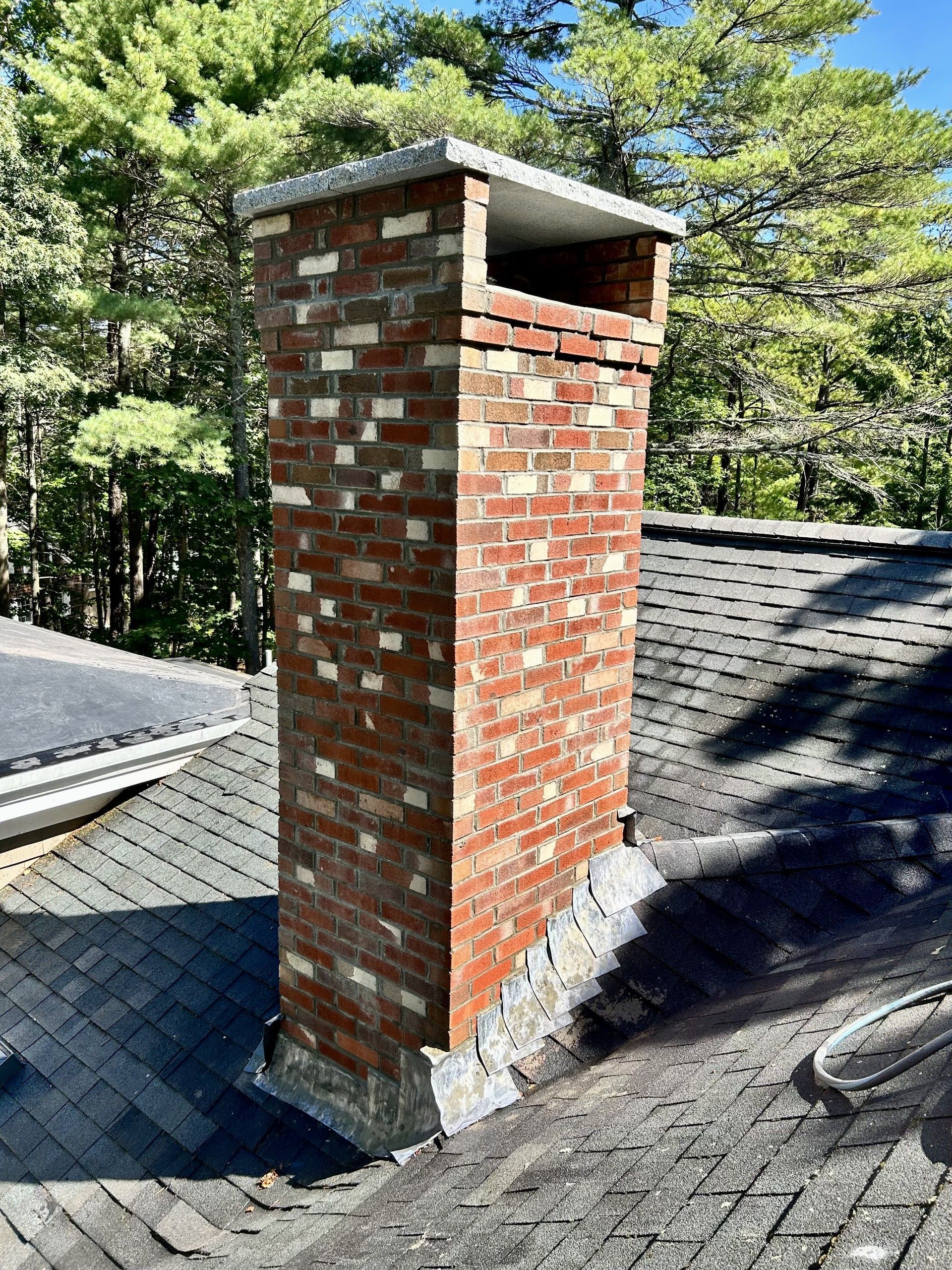 A red brick chimney with a gray stone cap on a shingled roof, surrounded by trees on a sunny day.