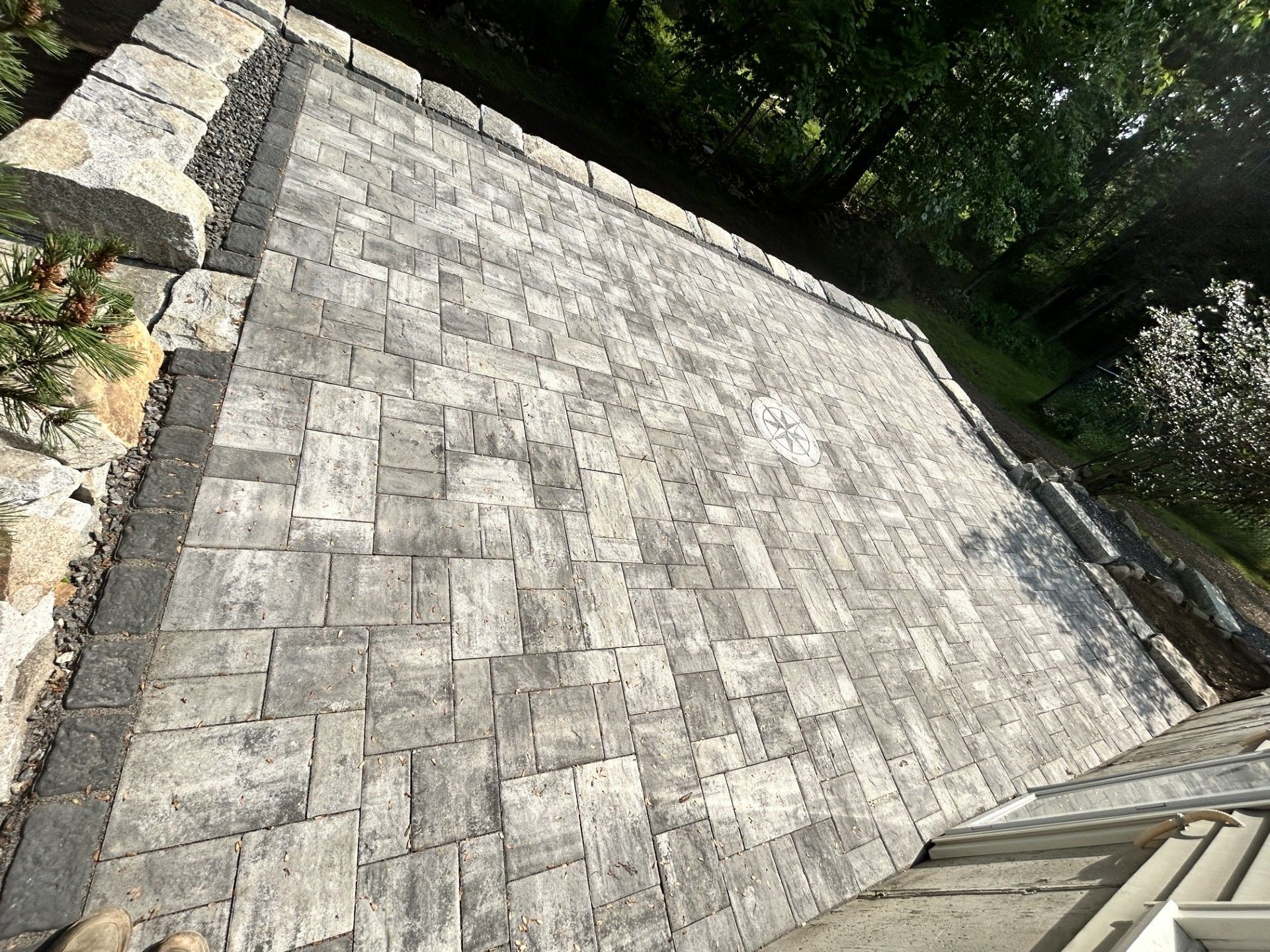 A newly installed gray paver stone patio featuring a darker decorative border, set next to a stone wall and landscaping.