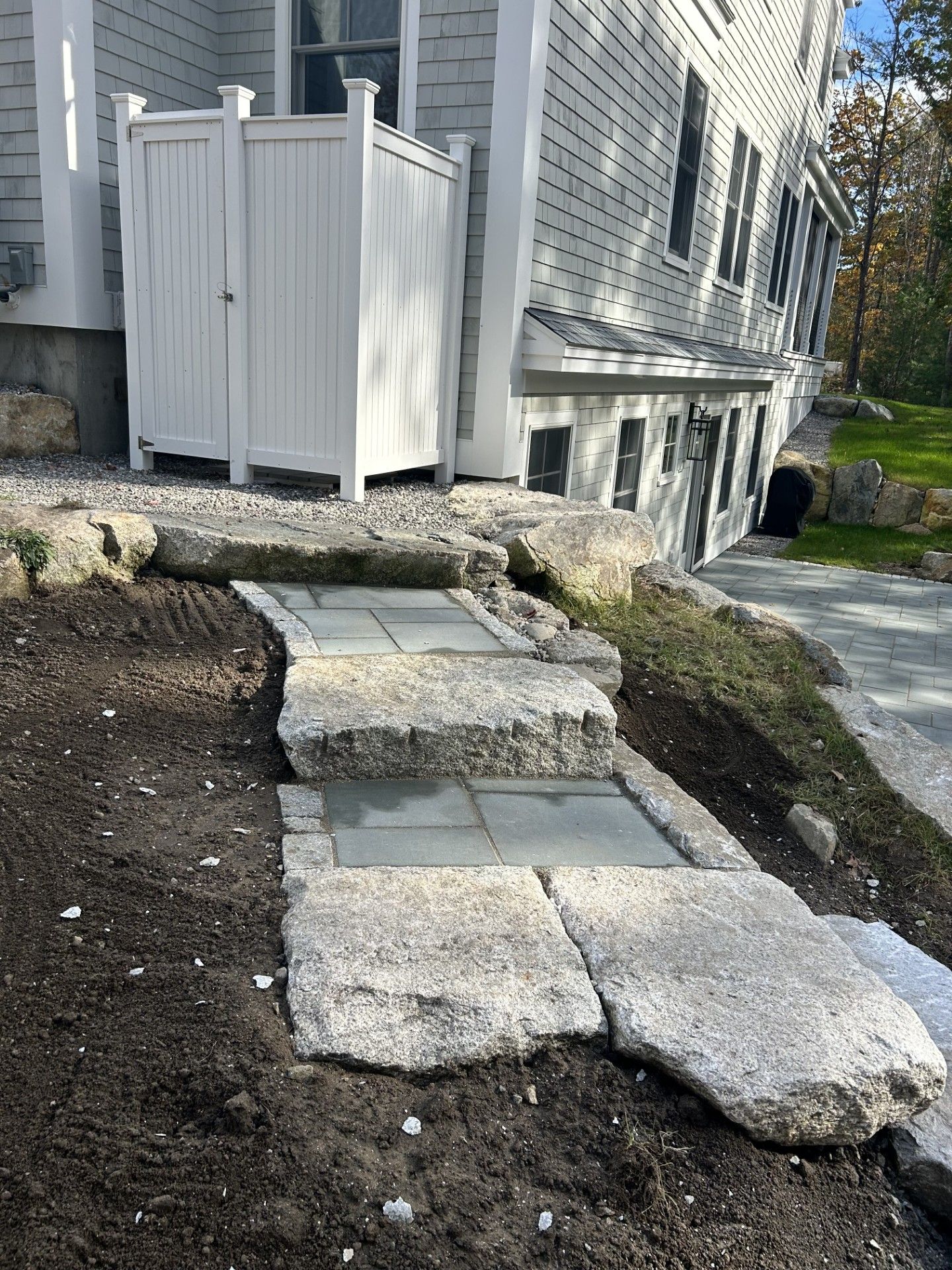 A path of large, rough-hewn stone steps interspersed with grey pavers leading up to a white fence near a house exterior.