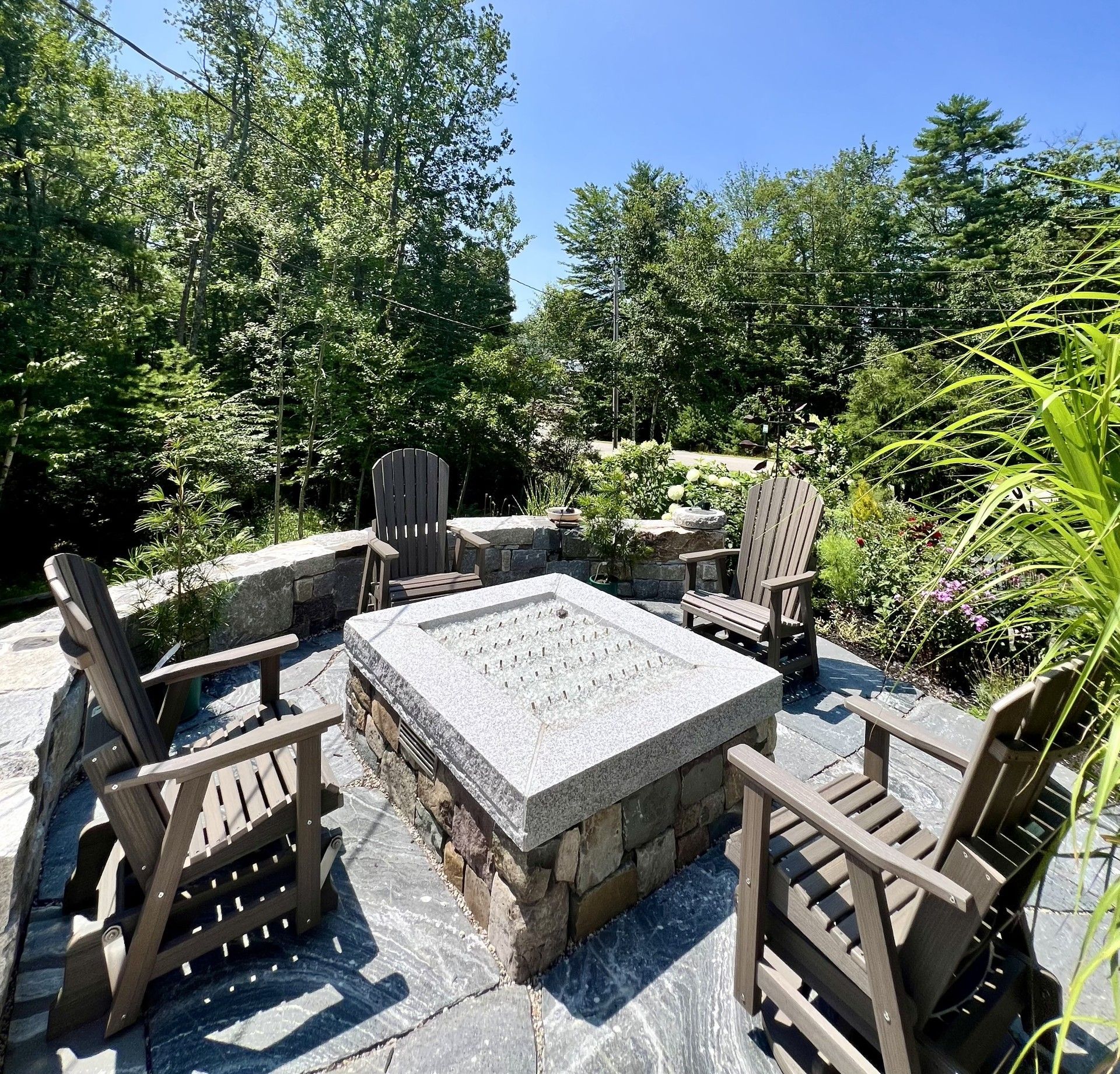 A granite stone fire pit sits on a slate patio surrounded by four brown Adirondack chairs in a sunlit garden.