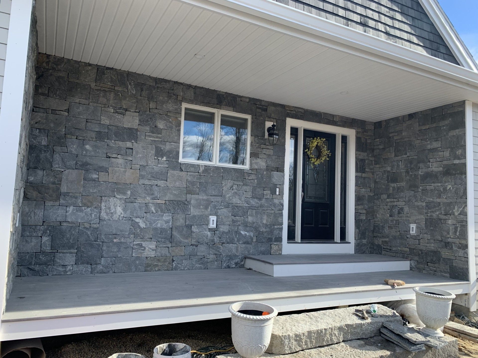 A front porch with grey stone walls, a dark door with a wreath, a white ceiling, and concrete steps with white planters.