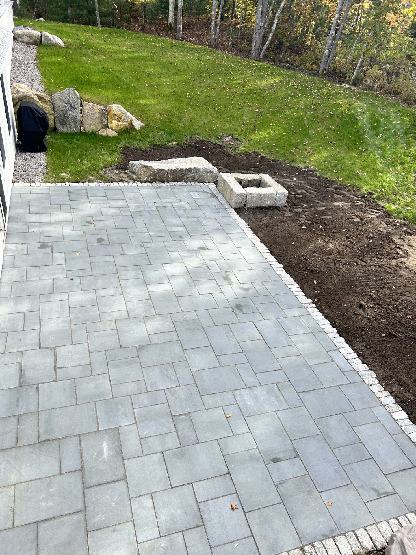 A grey rectangular stone patio, bordered by pavers, next to a patch of dark soil and a sloped grassy lawn with rocks.