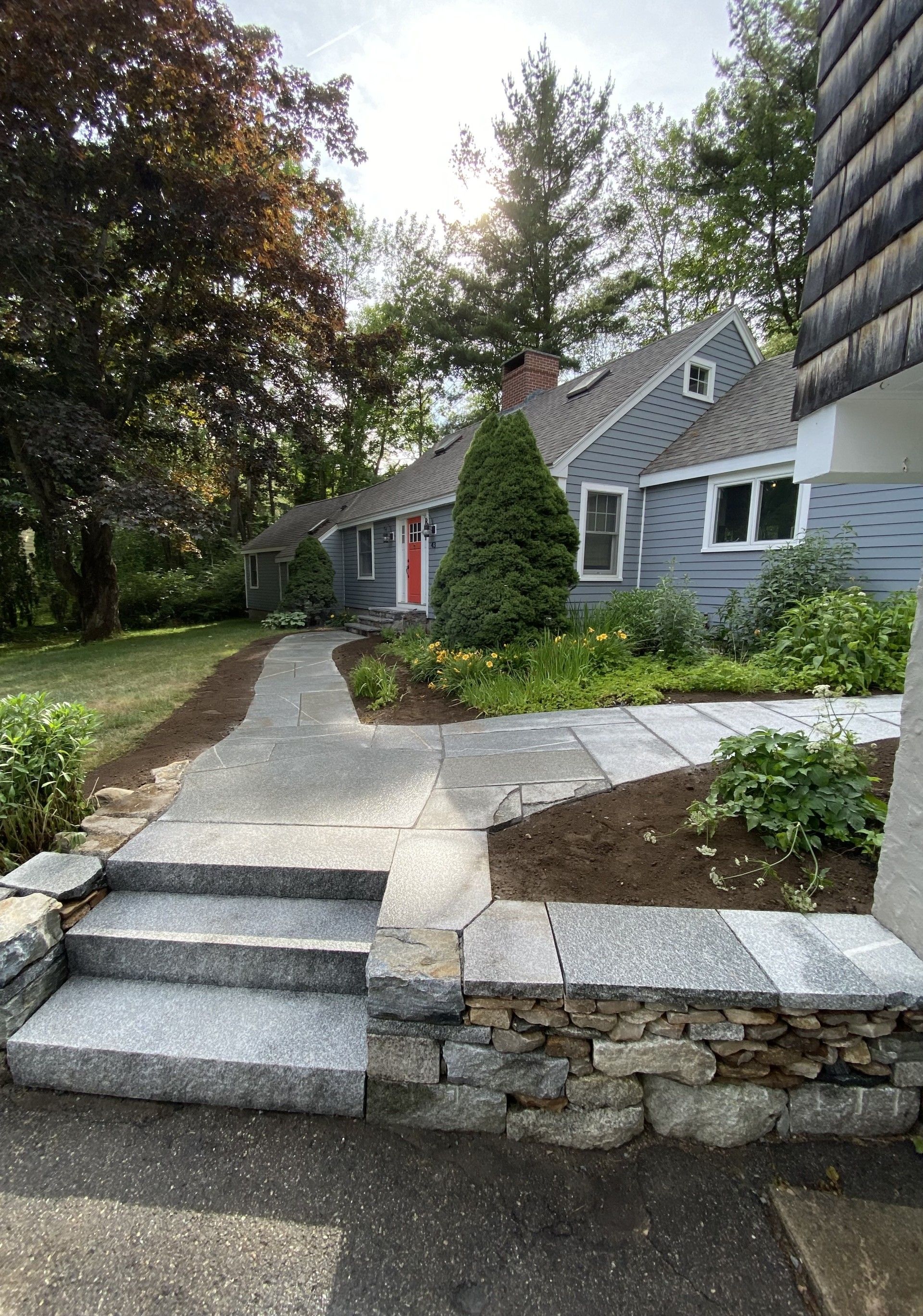 Stone steps lead to a walkway and a house with blue siding, a red front door, and surrounding landscaping.