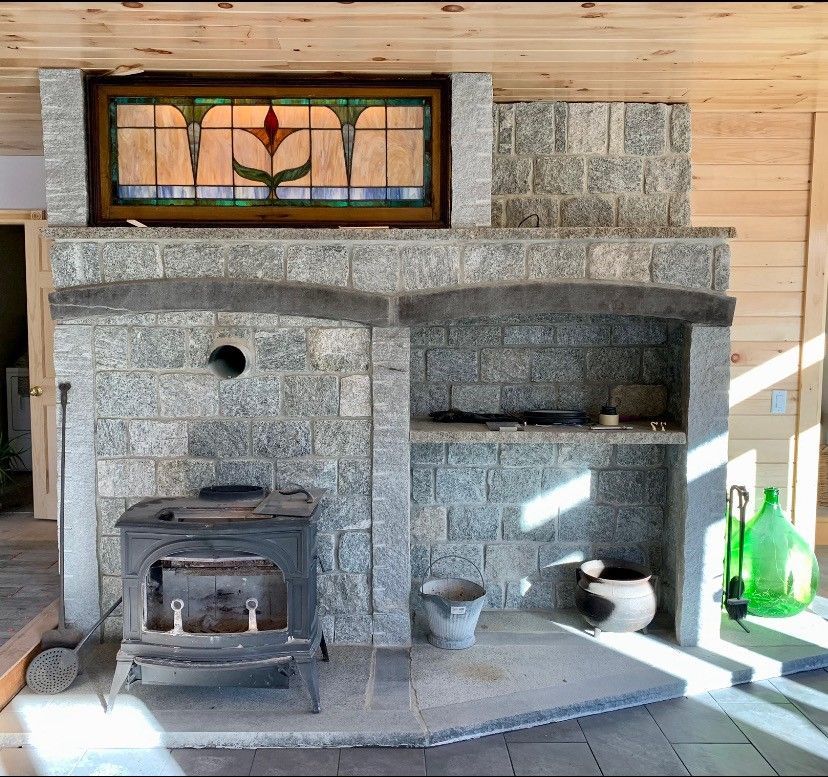 A rustic gray stone fireplace with a wood-burning stove on the left, a stone shelf on the right, and stained-glass inlay.