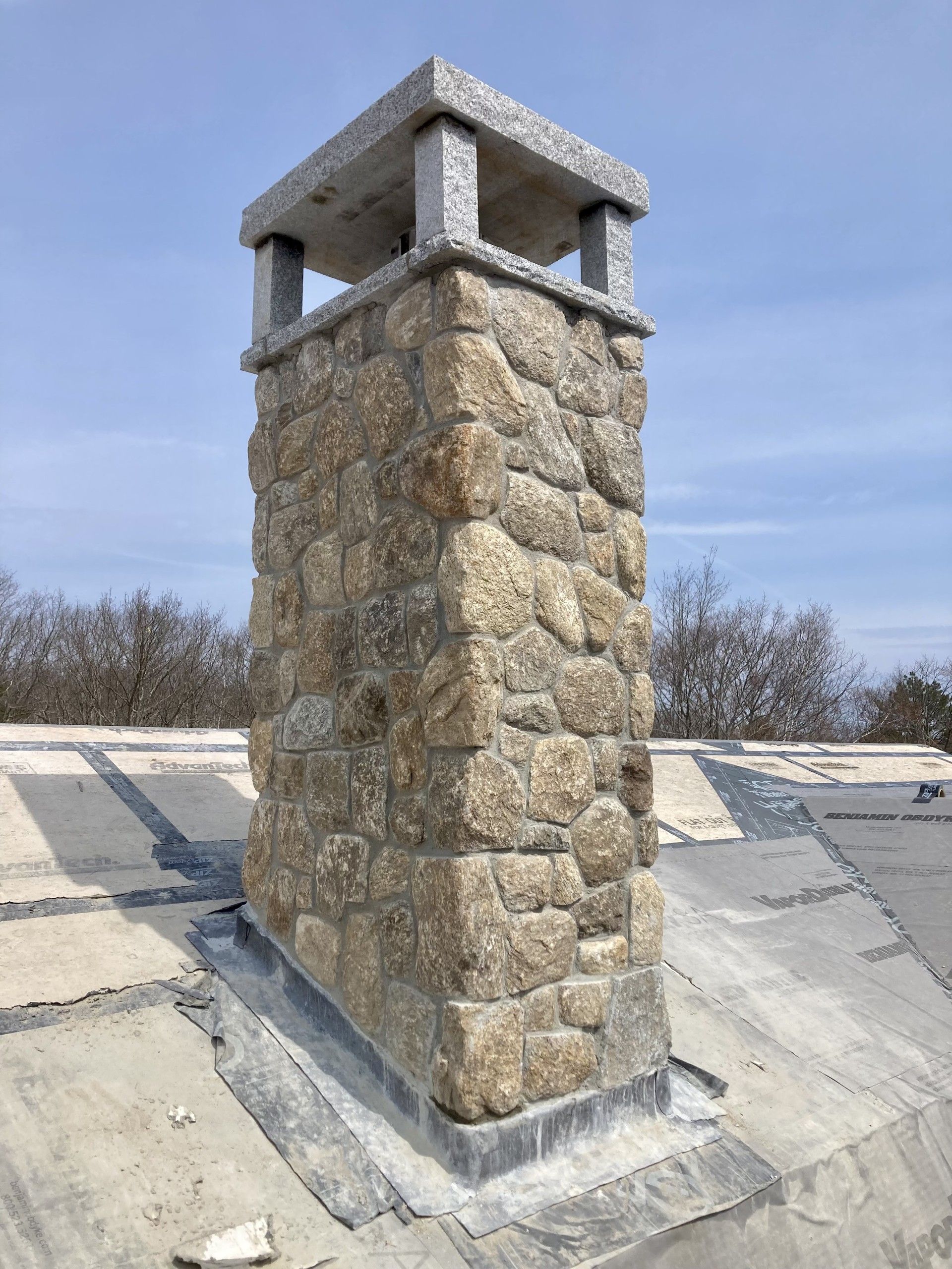 A stone chimney with a concrete cap sits on a grey, weathered roof against a clear blue sky.