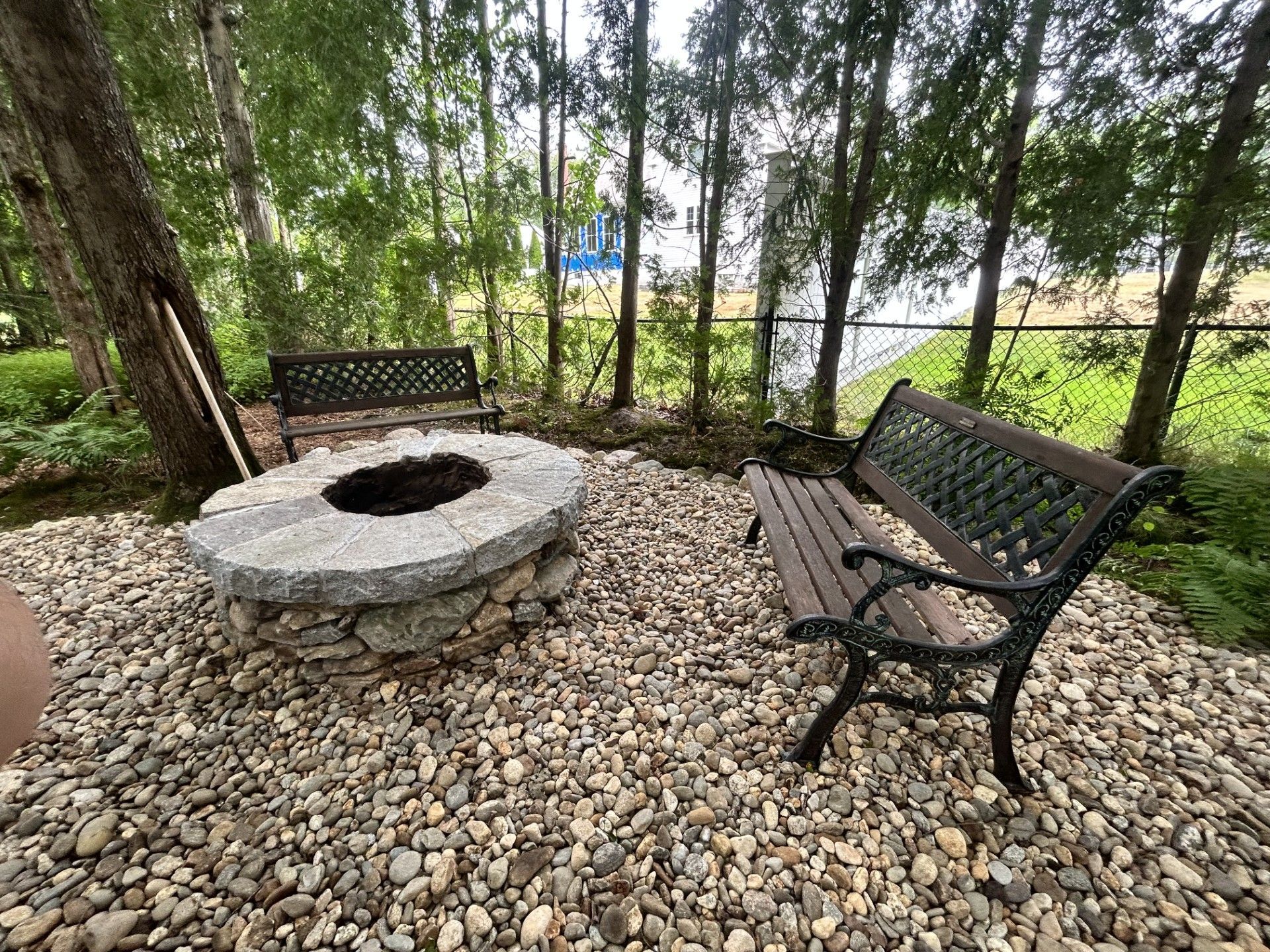 A stone fire pit surrounded by gravel and two dark metal park benches in a wooded backyard.