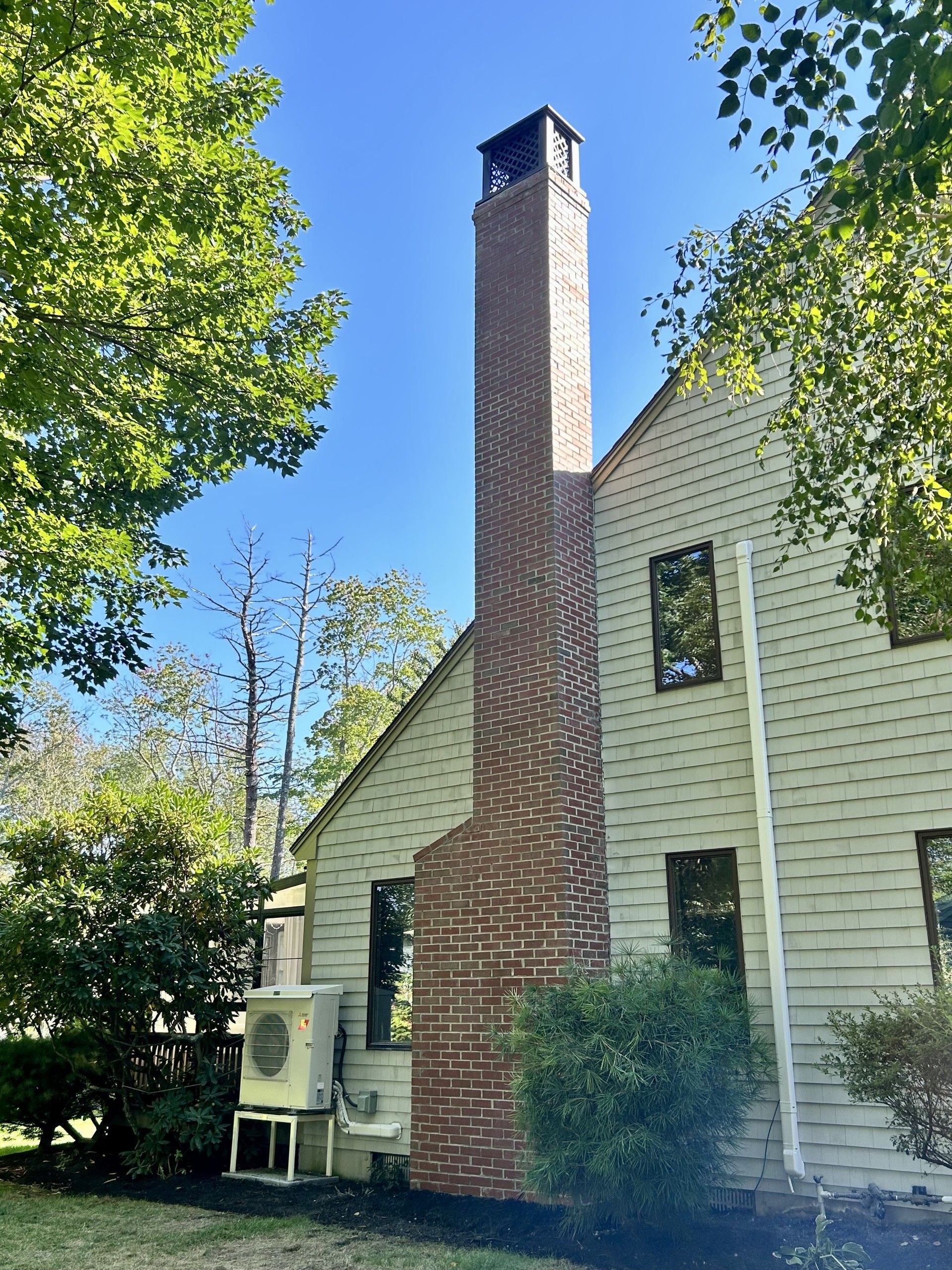 Exterior view of a house with a tall brick chimney, beige siding, and an air conditioning unit on the ground.