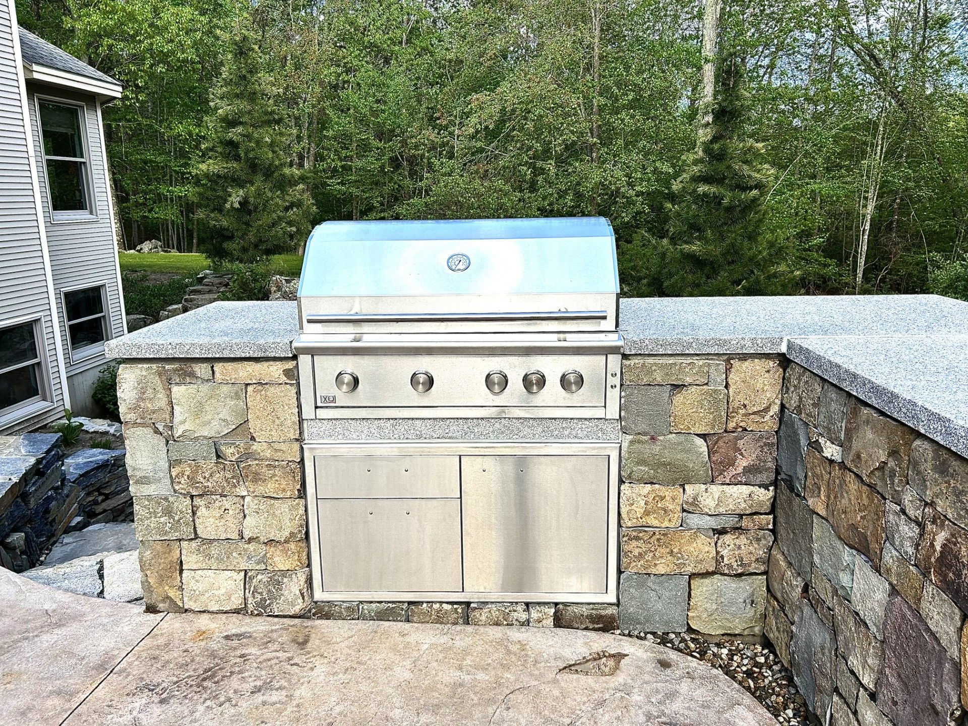 A stainless steel outdoor grill built into a rustic stone kitchen island on a patio with trees in the background.