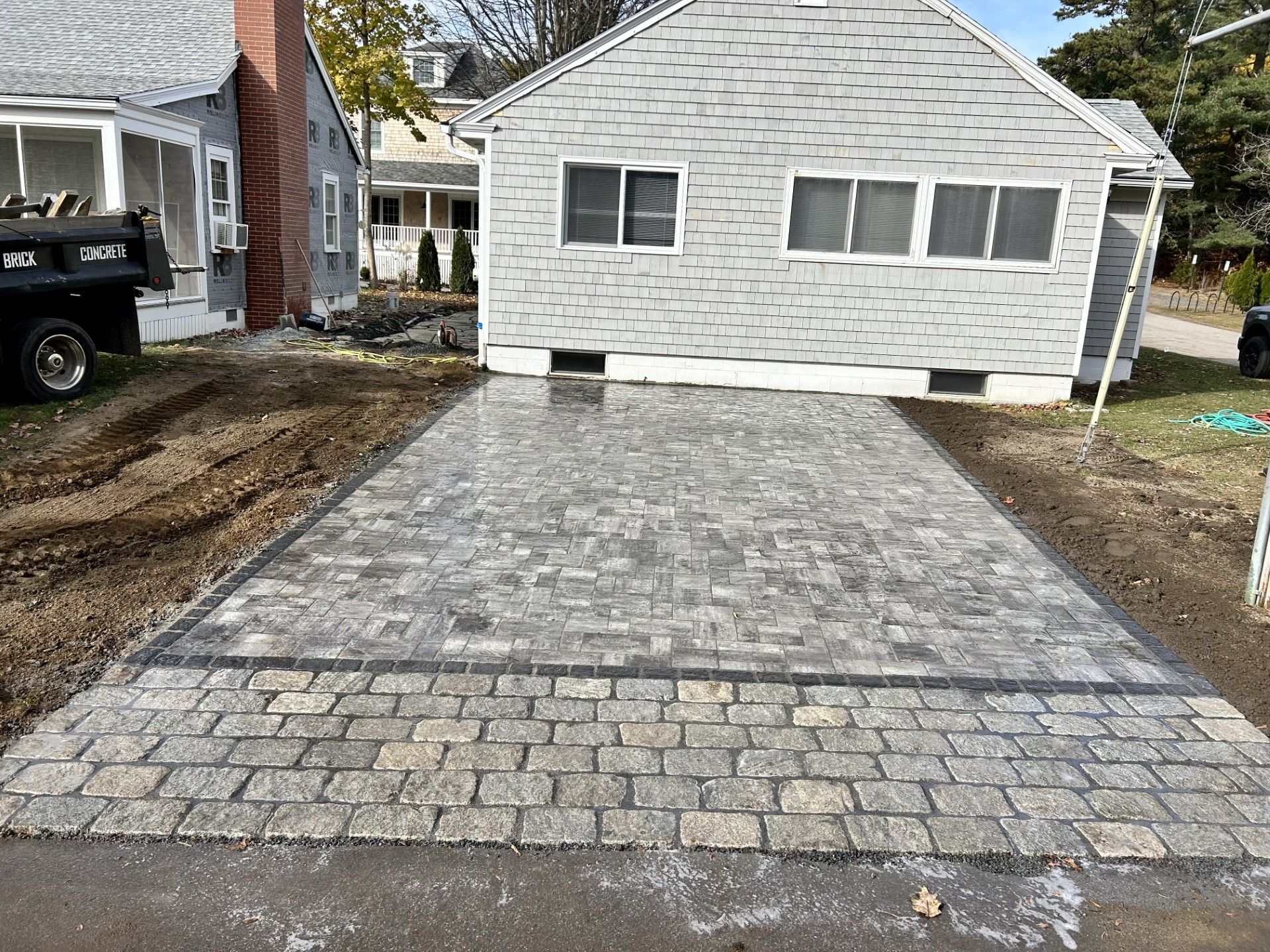 A paved stone patio transitioning into a walkway in front of a house with grey siding.