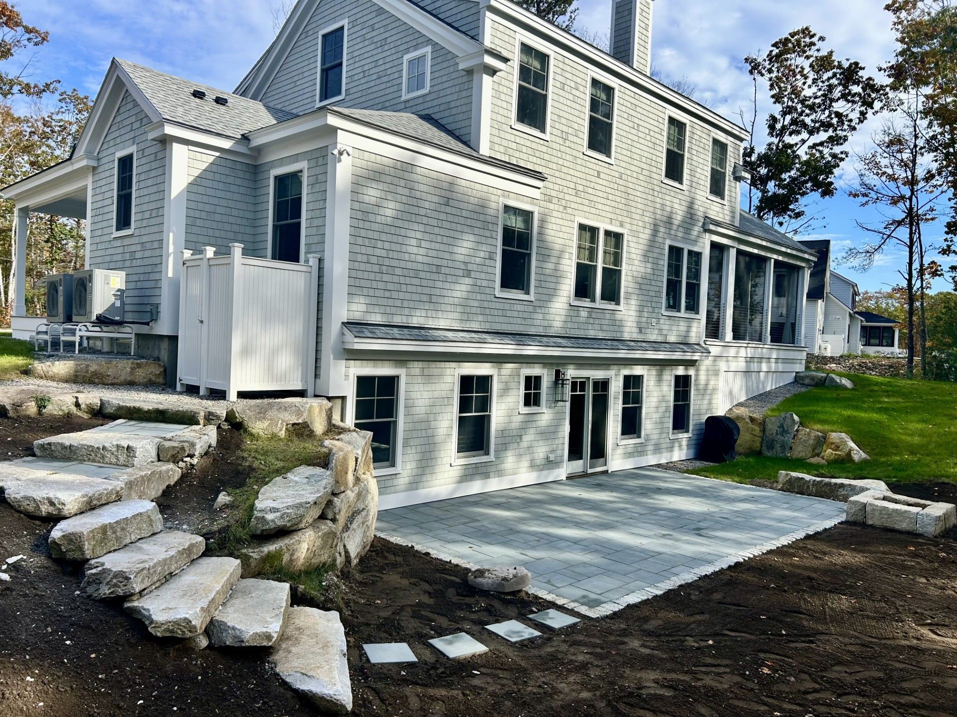 A light grey shingled two-story house with a stone staircase leading to a ground-level patio and a landscaped yard.