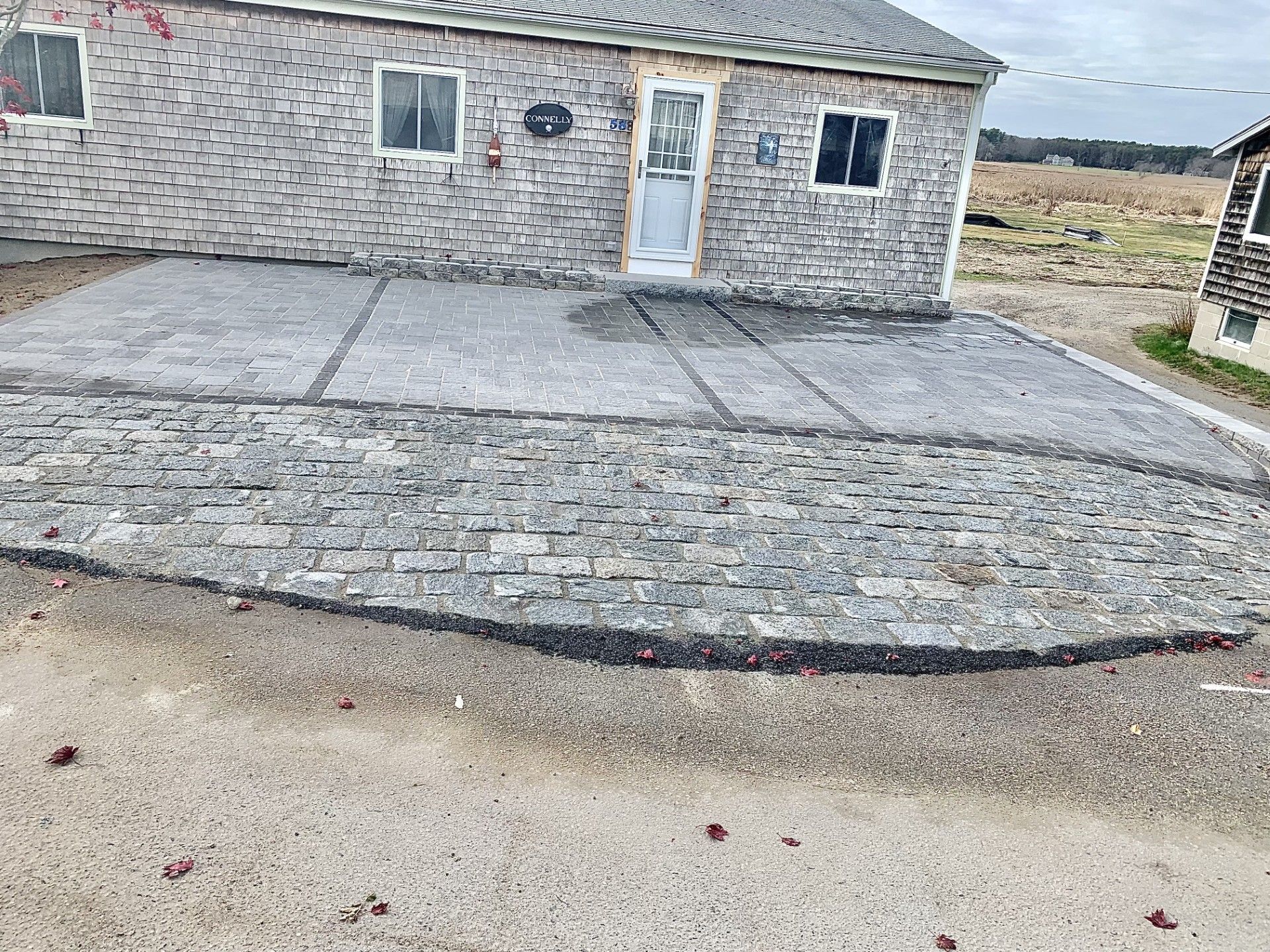 A newly installed stone patio and concrete walkway leading to the front door of a light-colored brick house.