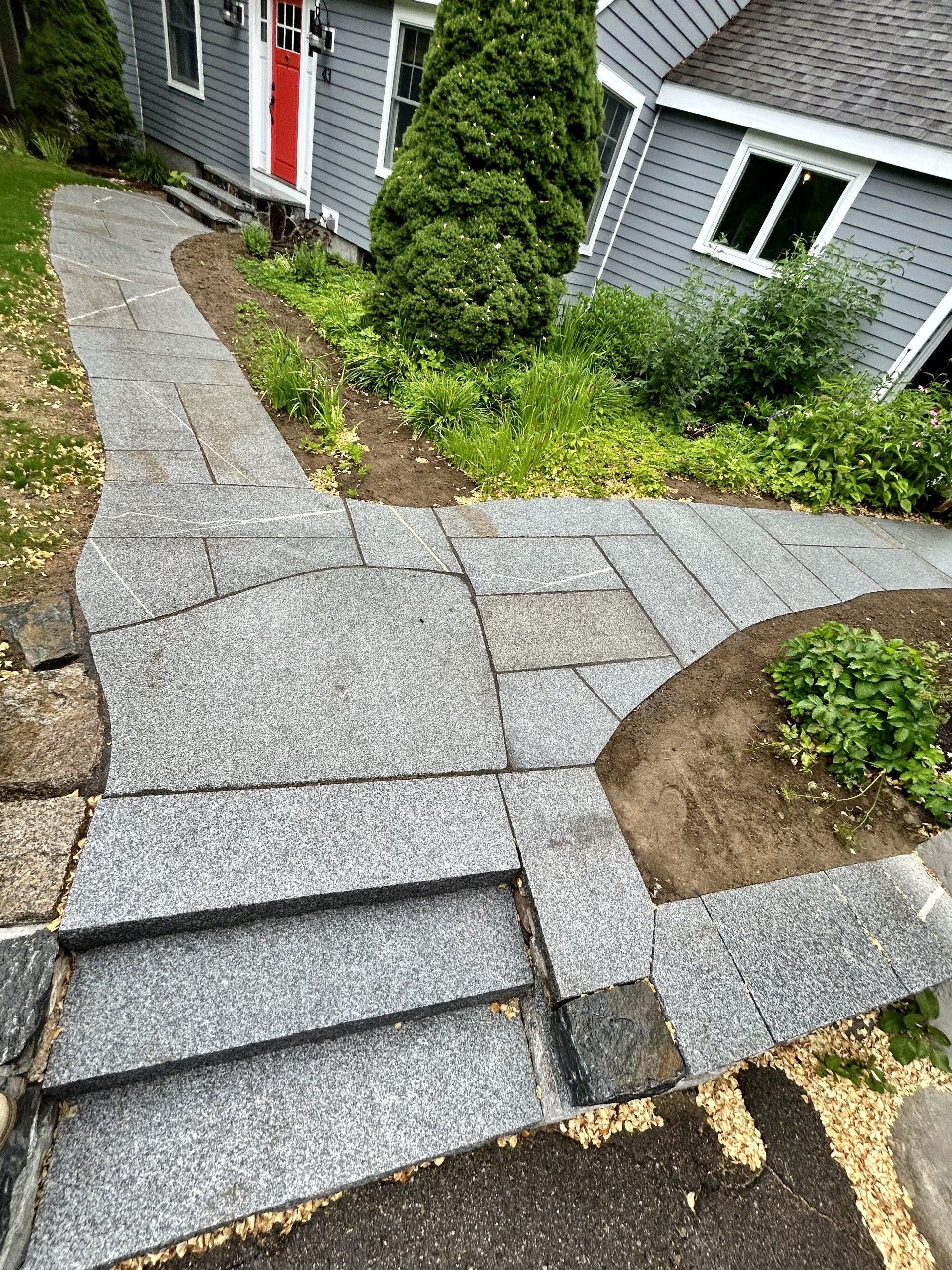 A granite paved walkway with steps leads to a house with grey siding and a red front door.