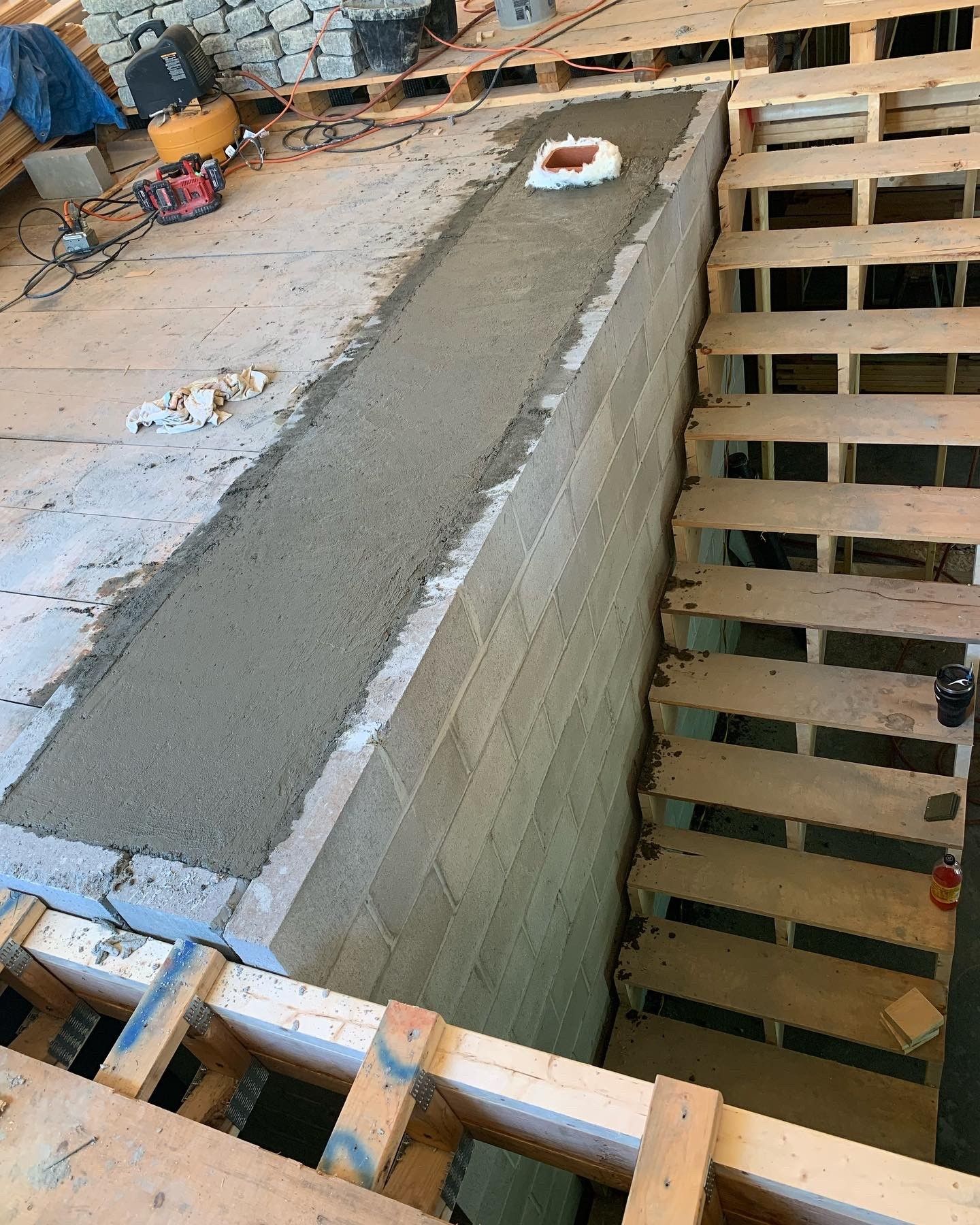 A construction site view of a block wall topped with wet mortar, adjacent to wooden stairs under framing.