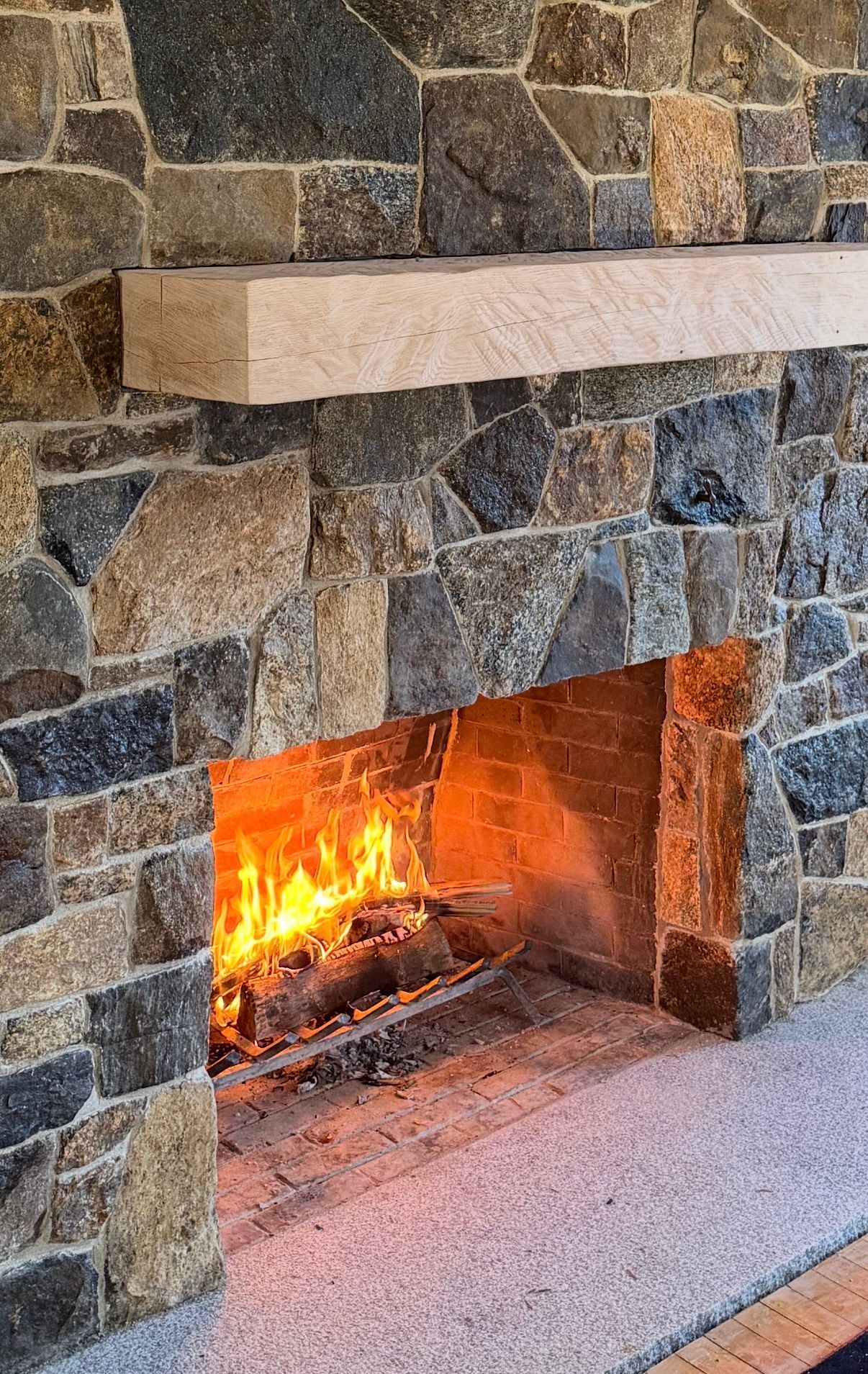 A cylindrical fire pit made of stacked natural stones centered on a gray paver patio in front of a white cottage.