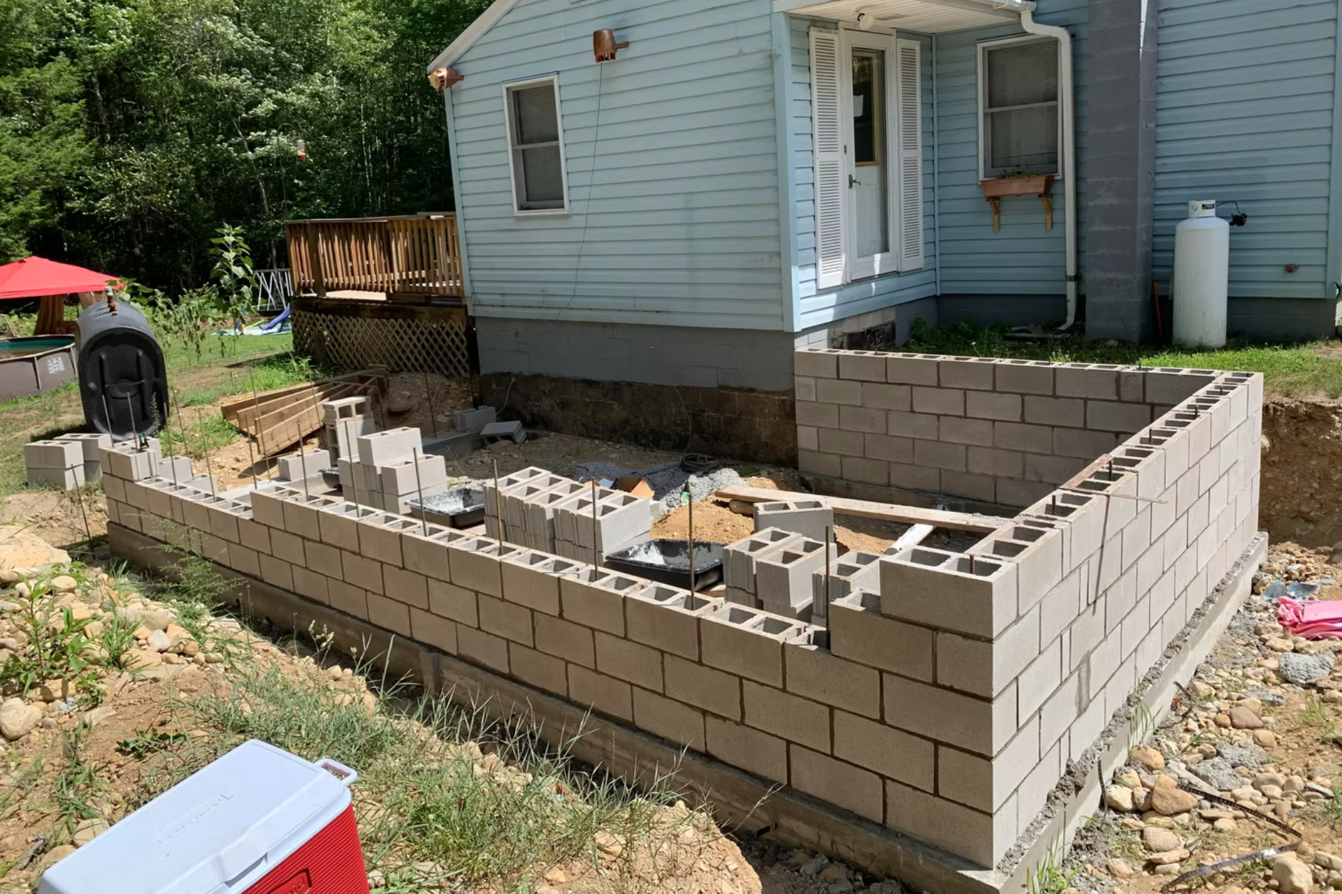A partially constructed concrete block wall foundation for a home addition, set in a backyard near a light blue house.