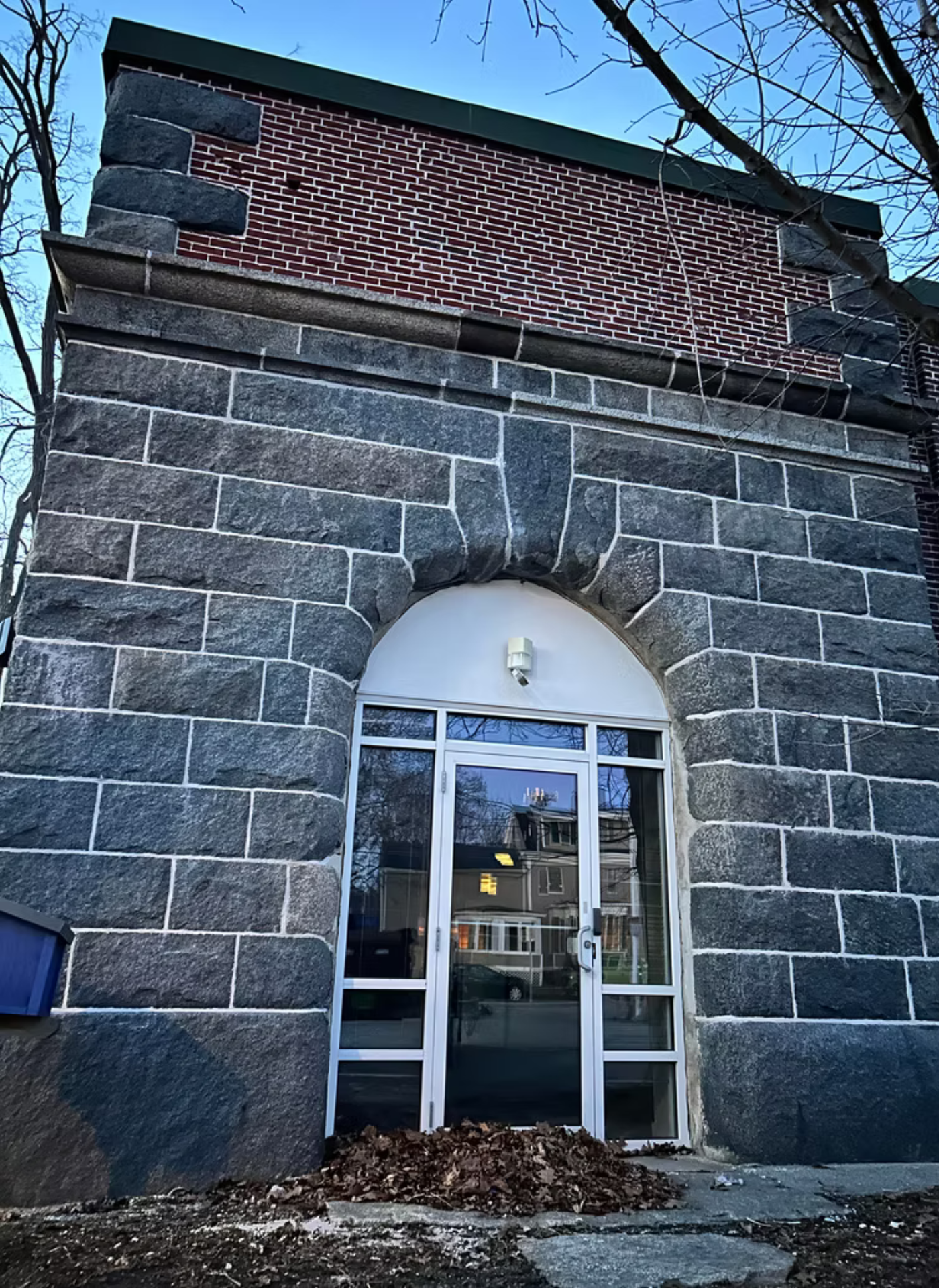 A stone building entrance with a glass door under a rounded arch, topped by a section of decorative red brickwork.