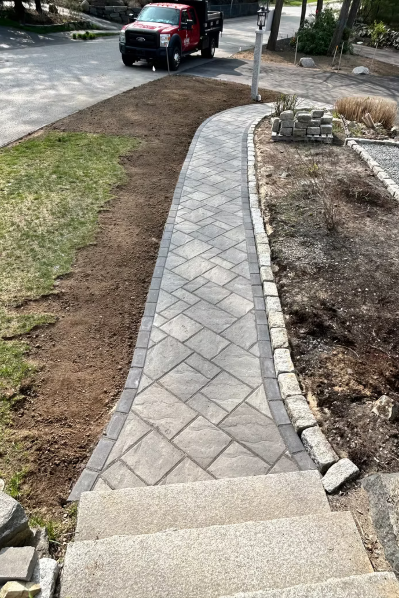 A stone walkway leads from concrete steps through a yard, bordered by gray pavers and dark soil, with a truck parked nearby.