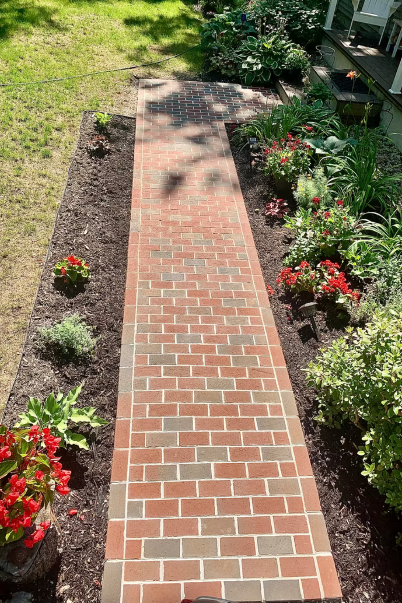 A straight brick walkway leading to a house, flanked by garden beds with dark mulch and blooming flowers.