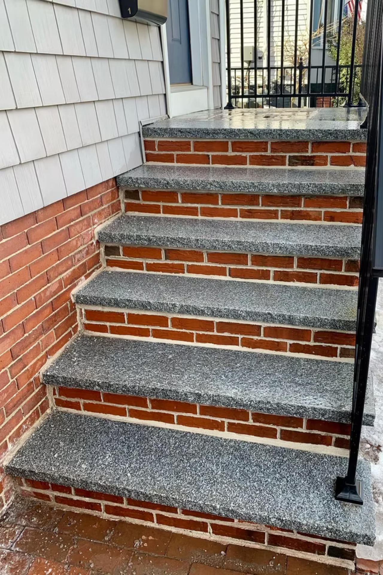 A flight of five brick steps with grey stone treads leading to a house entrance with a black metal railing.