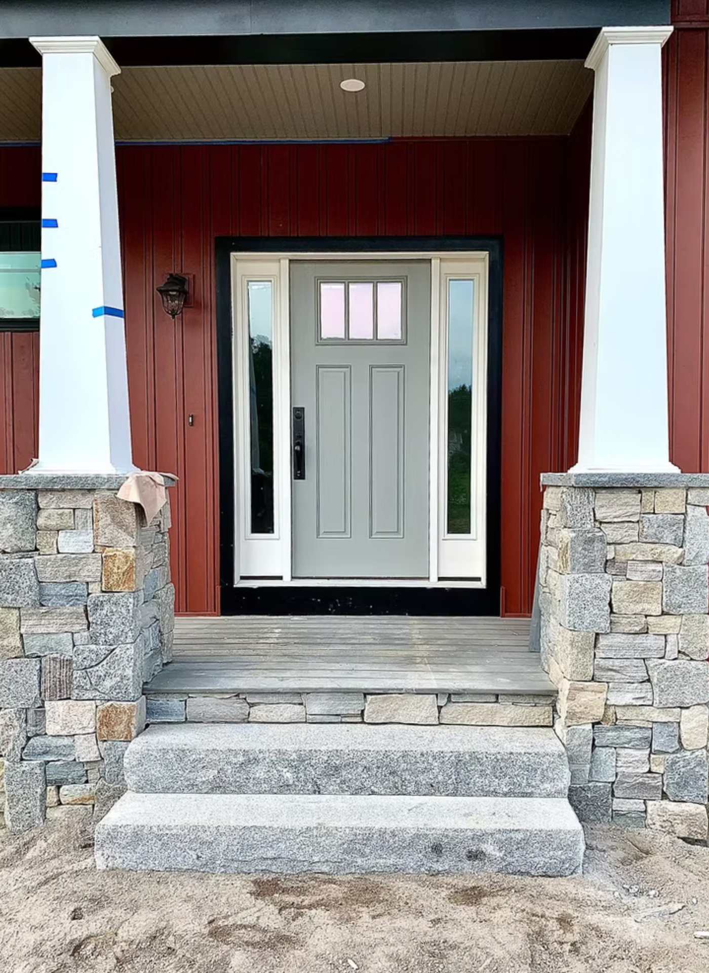 A grey front door with side windows between two white pillars on stone steps against a red metal exterior wall.
